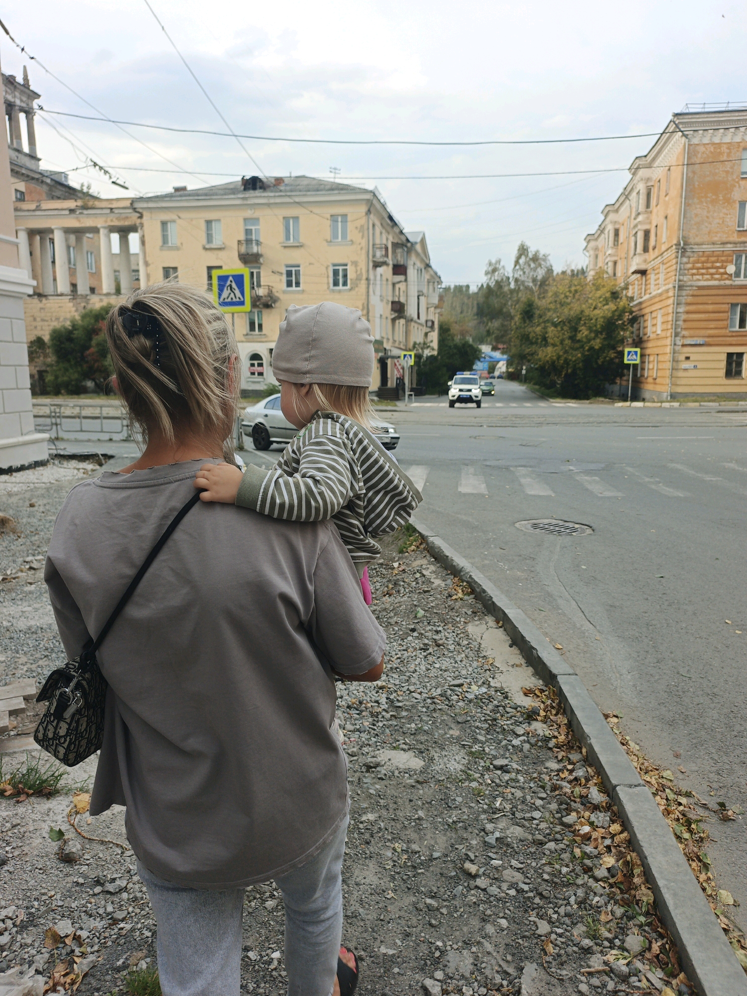 In Zlatoust, Russia, a woman with blonde hair pulled back carries a young child on her left shoulder. The woman wears a grey t-shirt and grey sweatpants, with a dark patterned bag slung across her body. The child, wearing a grey hat and a striped long-sleeved shirt, faces away from the camera and reaches a hand onto the woman's shoulder. They are positioned on a gravel sidewalk next to a paved road with a pedestrian crossing. In the background, yellow apartment buildings line the street, with trees and power lines visible under an overcast sky. Several cars are on the road, and traffic signs indicate a pedestrian crossing. The overall scene depicts a moment in an urban environment, with the woman and child seemingly walking or pausing near an intersection.
