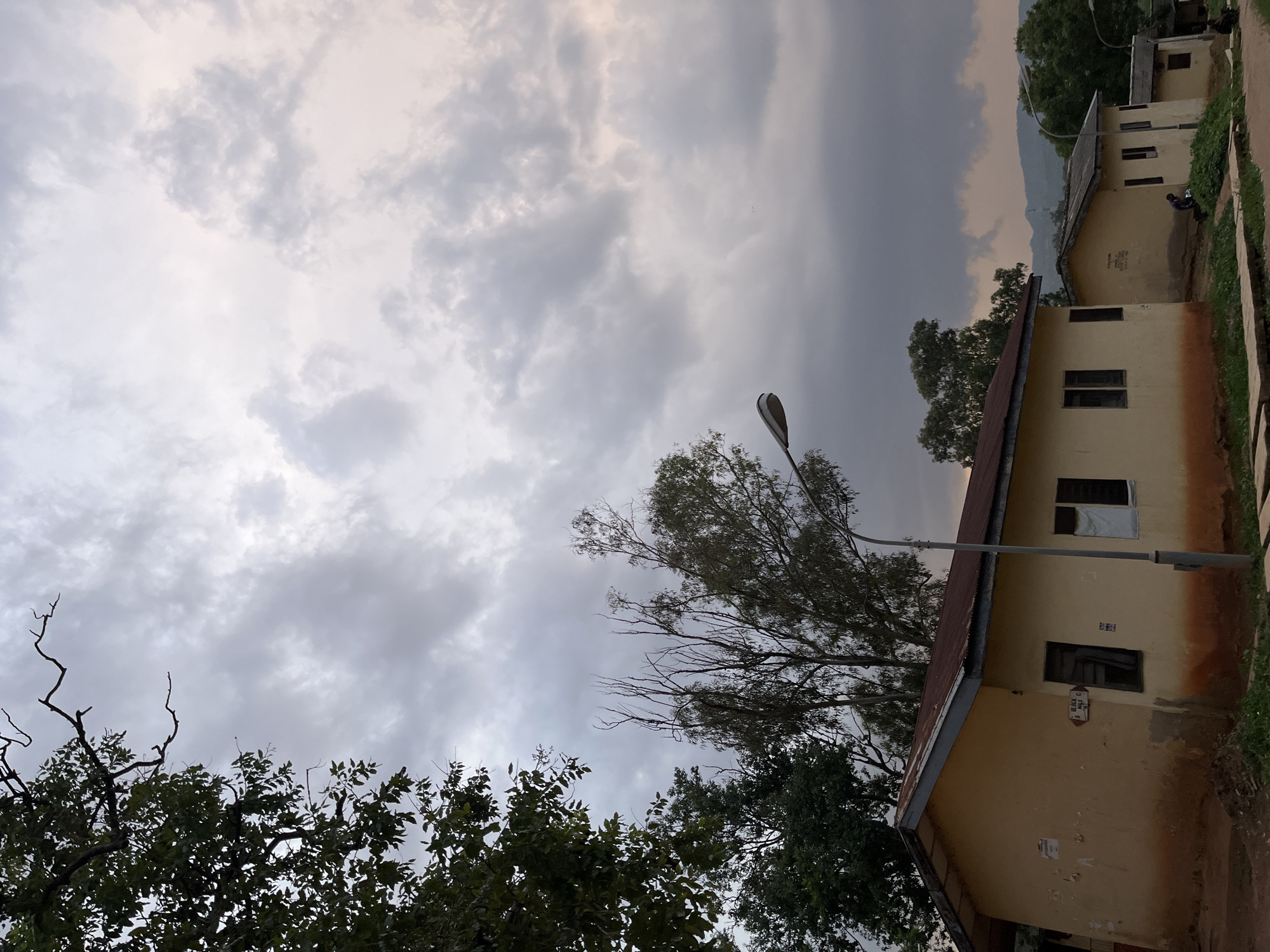 The image shows a street scene in Jos, Nigeria, under a cloudy sky. Two single-story buildings with yellow exteriors and reddish-brown roofs are prominent in the foreground. The building on the left has several windows and some faded posters or notices on its wall. A street lamp stands in front of it. The building on the right is similar in design, with a few windows and some vegetation growing in front. Further down the street, more buildings are visible, and in the background, hills can be seen against the overcast sky. A few individuals are present in the scene; one person appears to be tending to something near the building on the right, and others are seated further down the street. The environment appears to be an urban or semi-urban area, possibly a residential or institutional complex, given the simple architecture of the buildings and the presence of street lighting. The overall atmosphere is subdued, likely due to the cloudy weather.