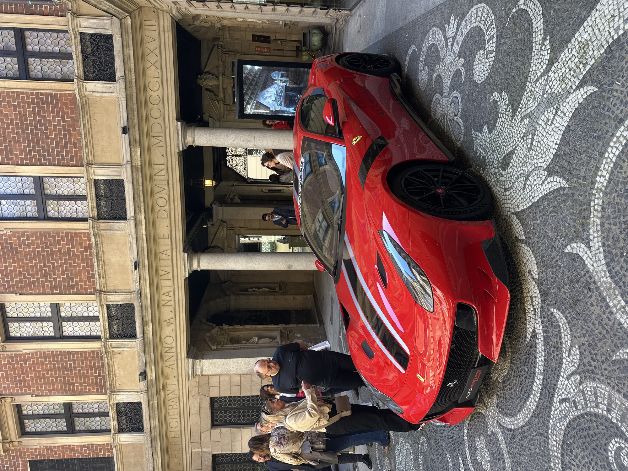 The image shows a red Ferrari parked in front of a historic building in Milan, Italy. The building has a stone facade with columns, inscribed with "T. PERFICIEBANT ANNO A. NATIVITATE DOMINI. MDCCLXXVII". The car is parked on a patterned stone pavement. Several people are standing around the car, looking at it. The building has multiple windows with decorative metal grills.
