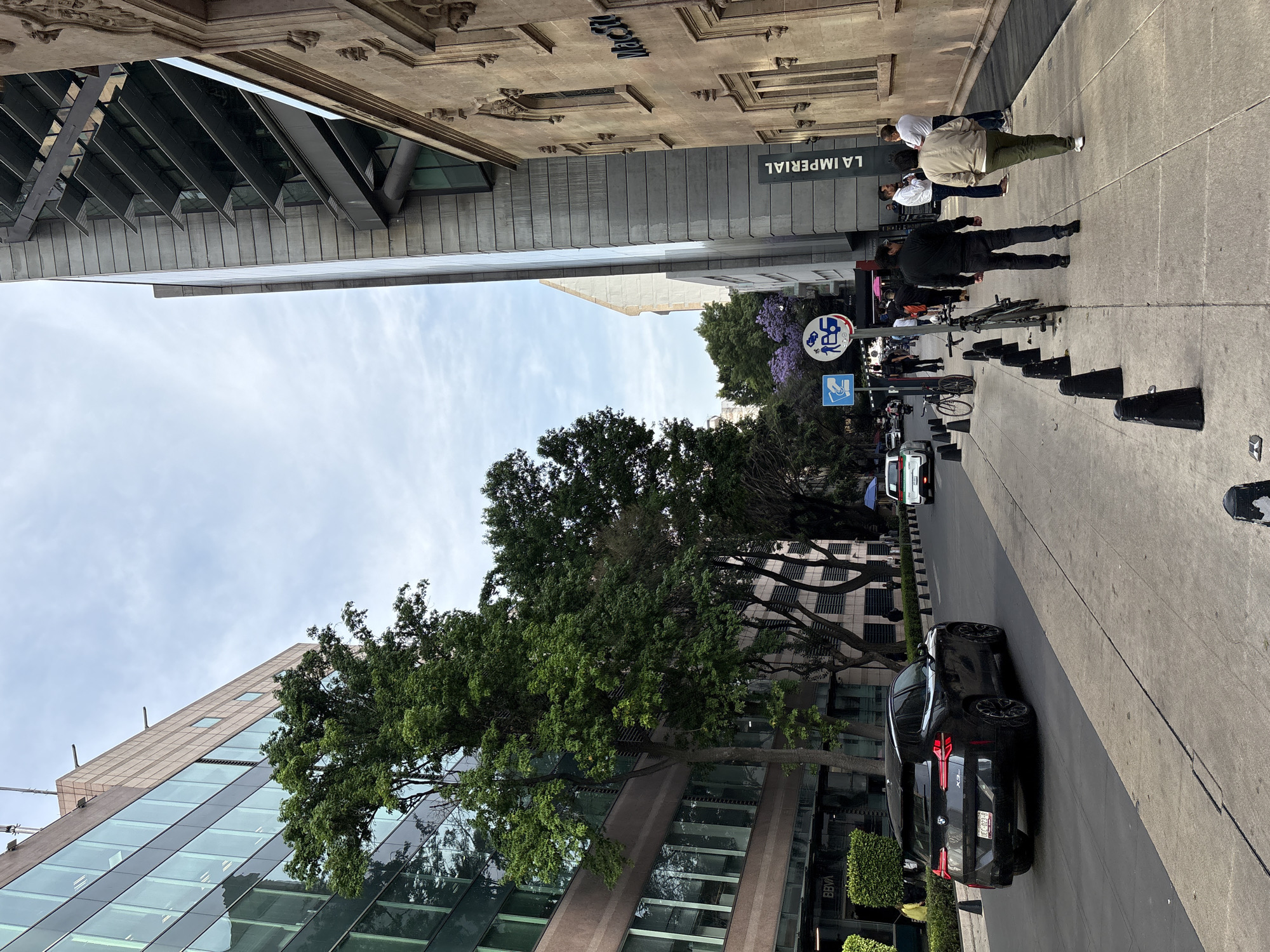 A street scene in Mexico City shows a busy sidewalk with pedestrians near the "La Imperial" building, identifiable by its signage. Cars are parked along the curb. Trees line the street. A sign depicting a bus indicates this is a bus stop. The sky is partly cloudy. The angle suggests the photograph was taken from a slightly elevated position. A modern glass-fronted building is visible on the left.