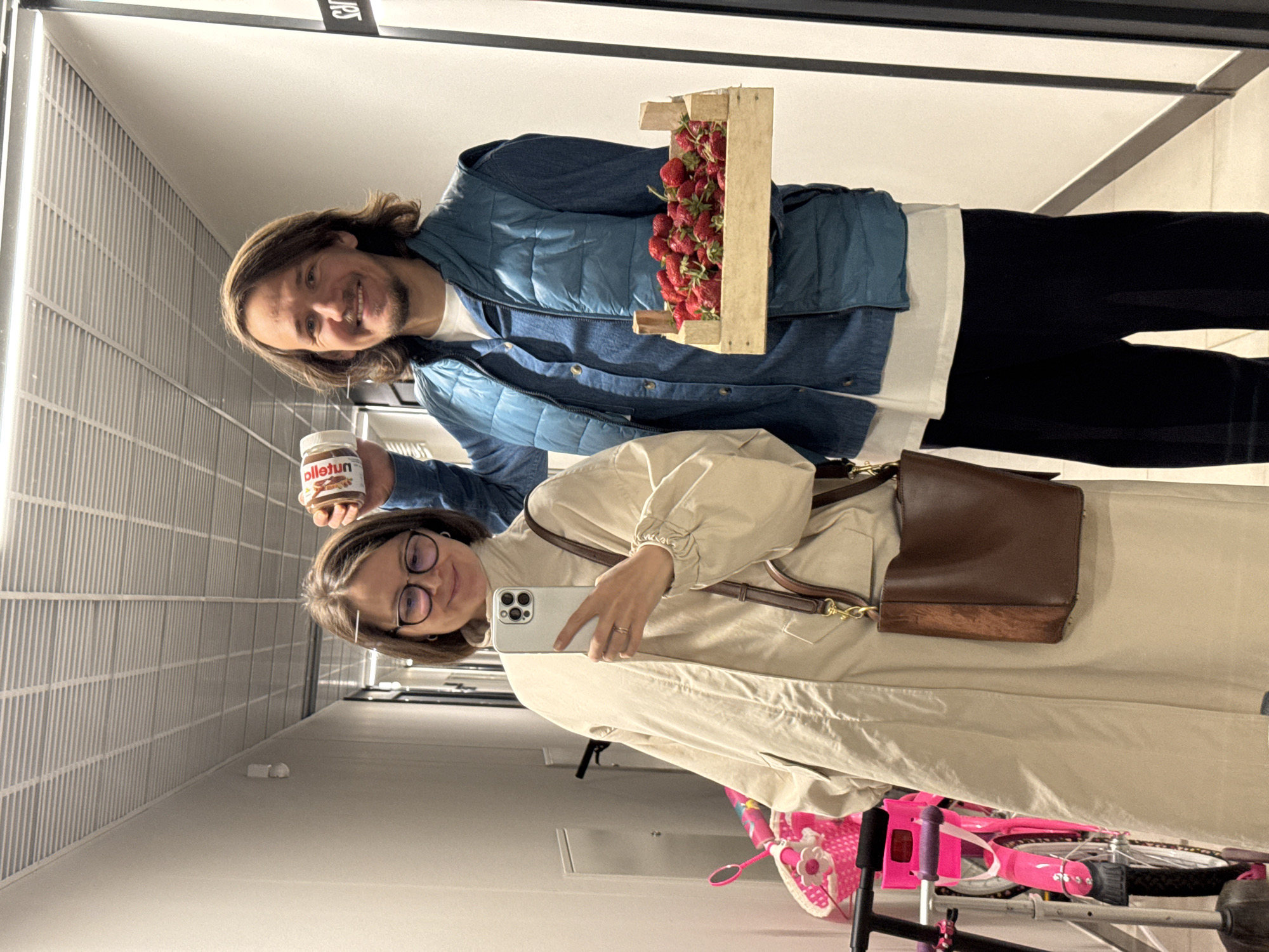 A waist-up shot depicts two people, a man and a woman, in a brightly lit interior hallway. The man is holding a wooden crate full of strawberries and a jar labeled "olletun." The woman holds a smartphone with multiple cameras (likely taking the picture), and has a brown purse with a shoulder strap hanging on her right shoulder. A pink children's bicycle with training wheels is partly visible in the bottom right corner. The interior has a modern design, with vertical striations in the wall, typical of the city Izhevsk, Russia.