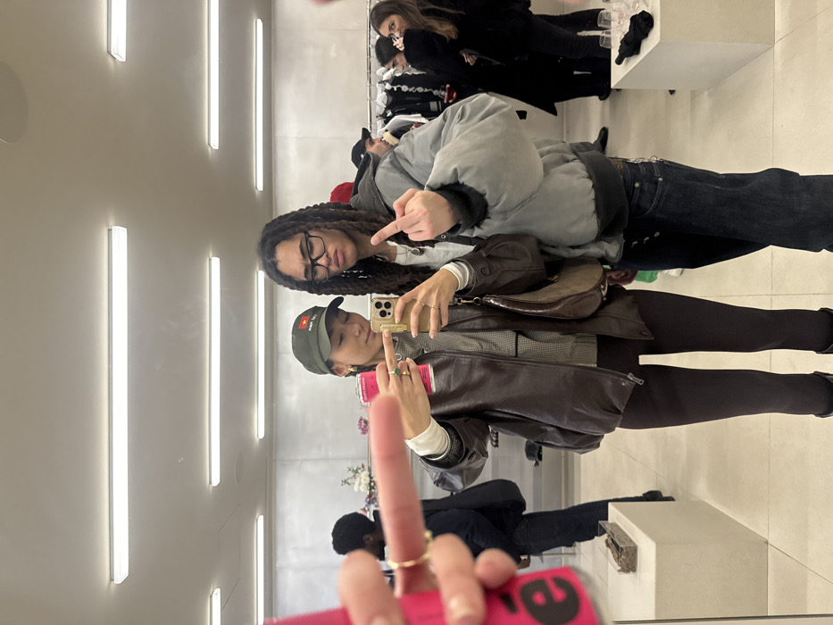Two people take a selfie in a mirror. One wears glasses and a grey puffer jacket, flipping off the camera. The other wears a green hat, brown leather jacket, and leggings, holding a phone with a pink case. Behind them are other people, clothing racks, and bright overhead lights. A man in a black suit sits in the mirror's reflection.