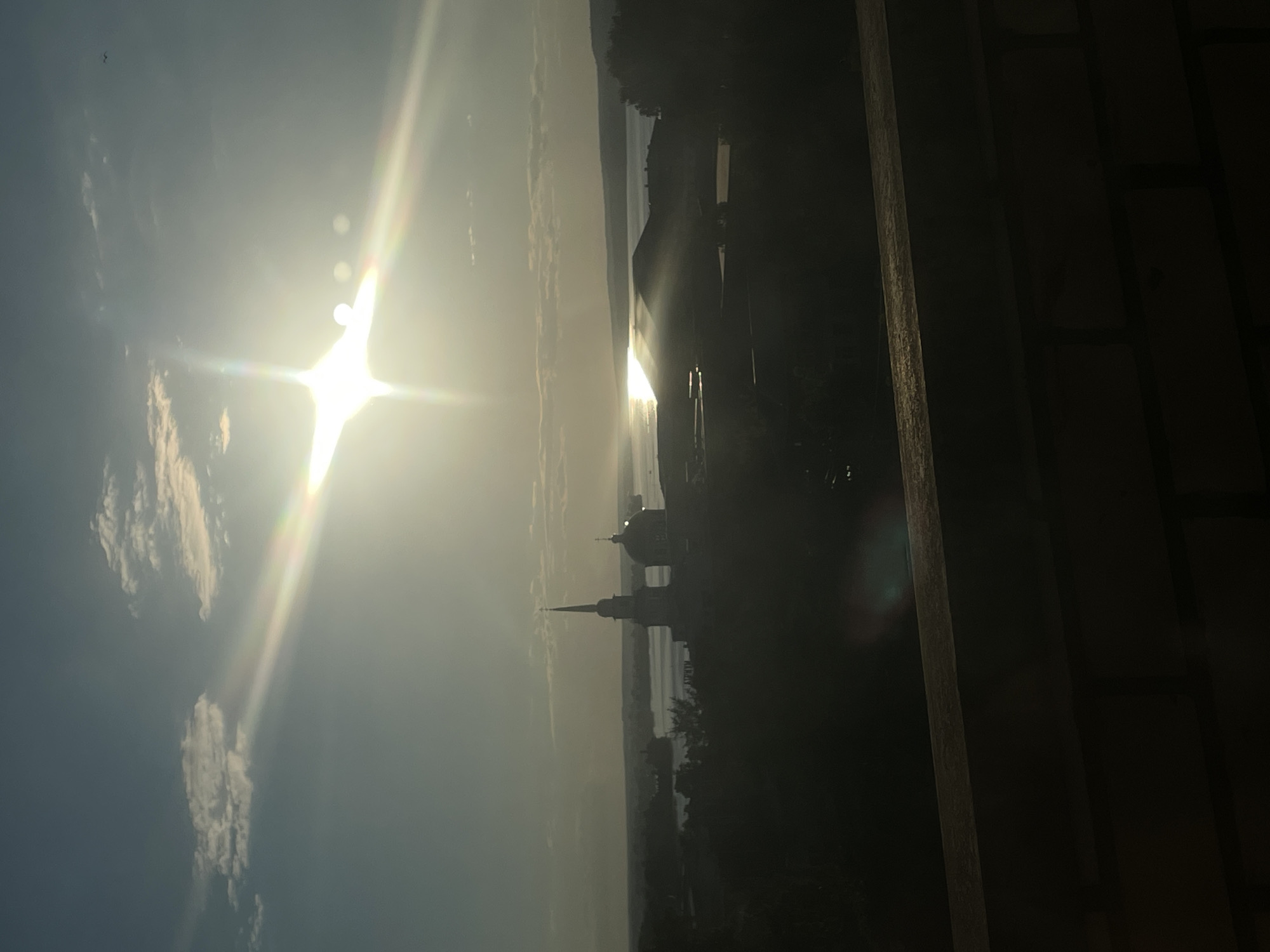 The image depicts a panoramic view at either sunrise or sunset, dominated by a brilliant sunburst and lens flare in the upper right quadrant of a hazy, light blue sky with scattered clouds. The sun's light reflects brightly off a wide body of water in the middle ground, which is characteristic of the Votkinsk Pond (Votkinsk Reservoir) in Votkinsk, Russia.
Silhouetted against the bright reflection and sky, in the mid-ground, stands a prominent architectural structure. This structure is identifiable as the Annunciation Cathedral (Blagoveshchensky Sobor) in Votkinsk, featuring a tall, slender spire on its left and a large, dark dome to its right. Other dark, indistinct buildings and tree lines are visible between the foreground and the water, and further along the distant shoreline. The immediate foreground shows a dark, indistinct brick wall or railing, suggesting the photograph was taken from an elevated vantage point. No people are discernible in the scene.