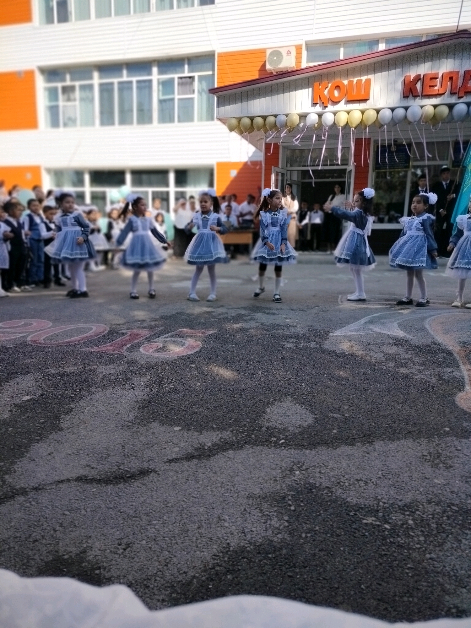 In Көлкент, Kazakhstan, a group of young girls dressed in blue school uniforms with white collars and bows are participating in an outdoor event, likely a school ceremony, in front of a building. The building features orange and white paneling, with large windows and a prominent orange sign above an entrance that reads "ҚОШ КЕЛДІ". Balloons are strung above the entrance. The ground in the foreground is asphalt, with the number "2015" written in chalk in a large font. Other students and adults are visible in the background, some standing and observing the event. The lighting suggests it is daytime, and the overall atmosphere appears festive.