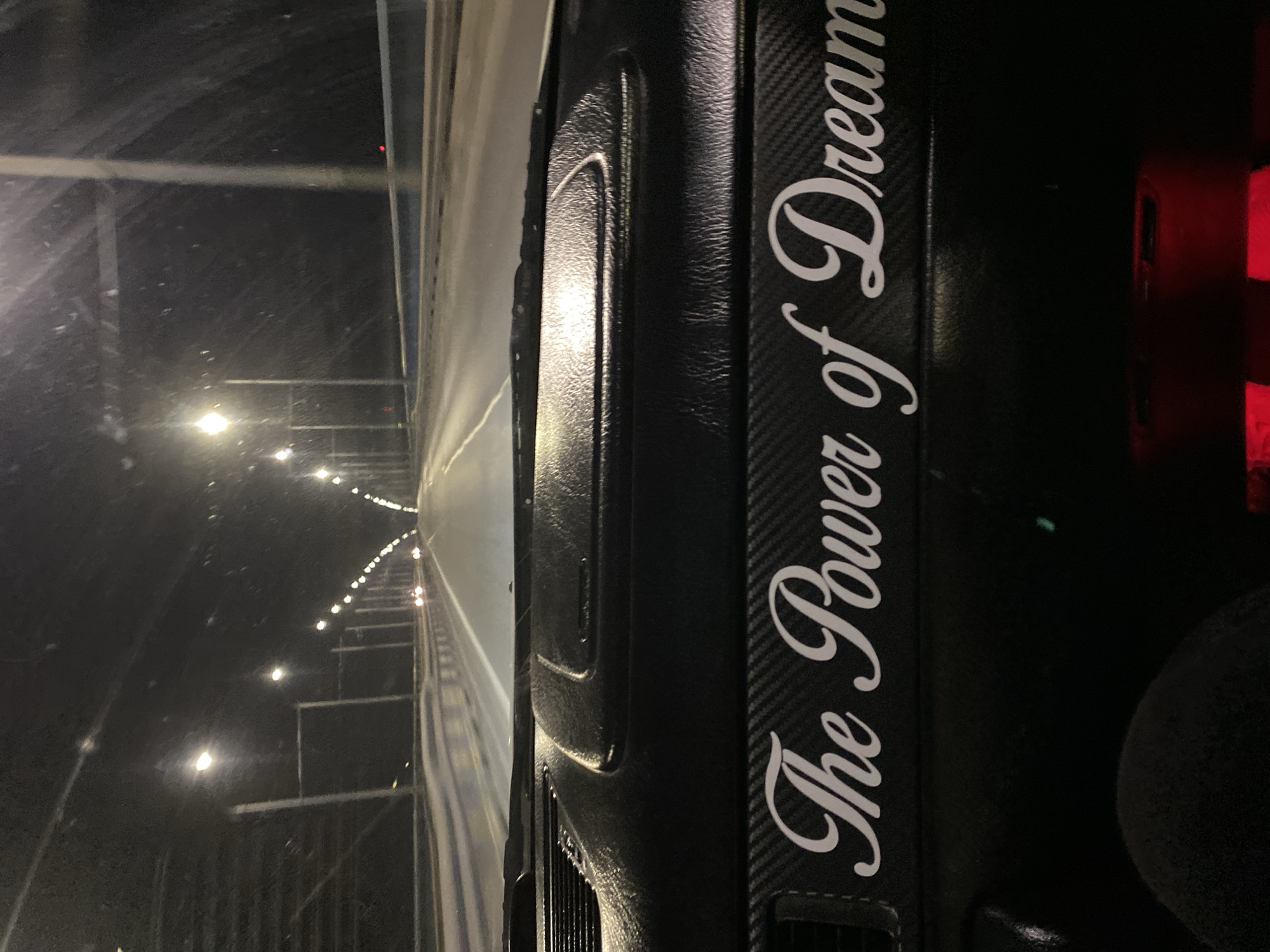 The image shows a view from inside a vehicle, driving through a tunnel. The tunnel is lit by evenly spaced lights on the sides. The dashboard of the vehicle is visible on the right side of the frame, with the words "The Power of Dreams" printed on it in white lettering. The background of the dashboard appears to have a carbon fiber pattern. Part of a steering wheel is also visible in the lower right corner. The scene likely takes place in the Asparuhovo bridge tunnel located in Varna, Bulgaria. No people are visible.