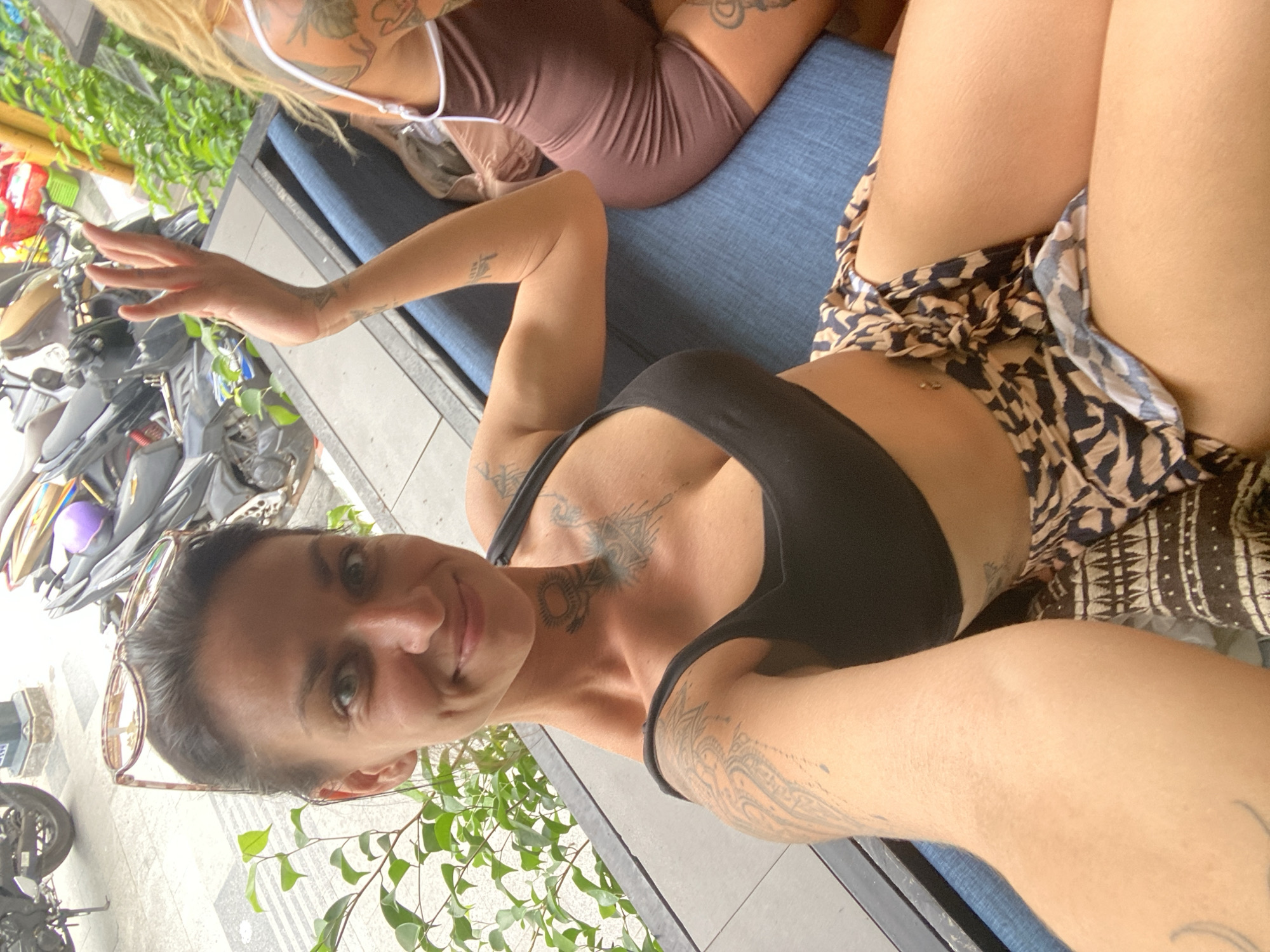 A woman is taking a selfie on a blue bench. She has dark hair and is wearing a black tank top and leopard print shorts. She is smiling and has tattoos on her arms and chest. Another woman with blonde hair is sitting next to her, wearing a brown leotard. The background shows a street with motorcycles and lush green plants. The scene appears to be in a city, possibly Phu Thuy Ward, Vietnam.
