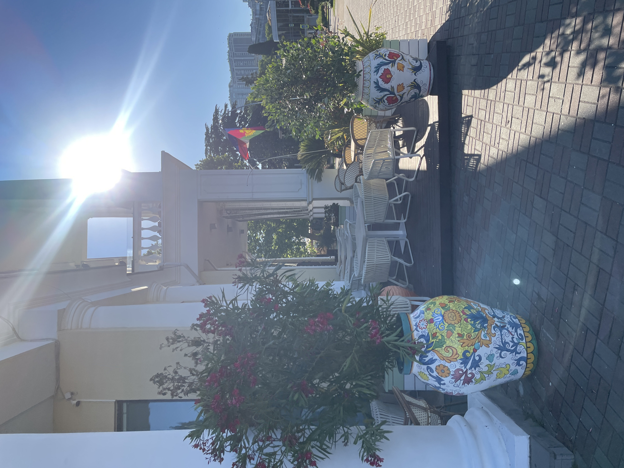 This image depicts an outdoor seating area of a cafe or restaurant located in the city of Sochi, Russia. The bright sun is shining intensely from the upper right corner of the frame, casting strong shadows. In the foreground, two large, ornate ceramic vases are prominently displayed. The vase on the left contains a lush green plant with pink flowers, while the vase on the right holds a smaller green plant. The seating area itself consists of several white tables and chairs, arranged on a dark wooden deck. Behind the seating area, a building with white columns and classical architectural elements is visible. A flag, featuring red, green, and yellow colors, is flying on a flagpole in the mid-ground. In the background, the cityscape of Sochi unfolds, with several tall buildings and trees under a clear blue sky. The ground in front of the seating area is paved with brickwork.