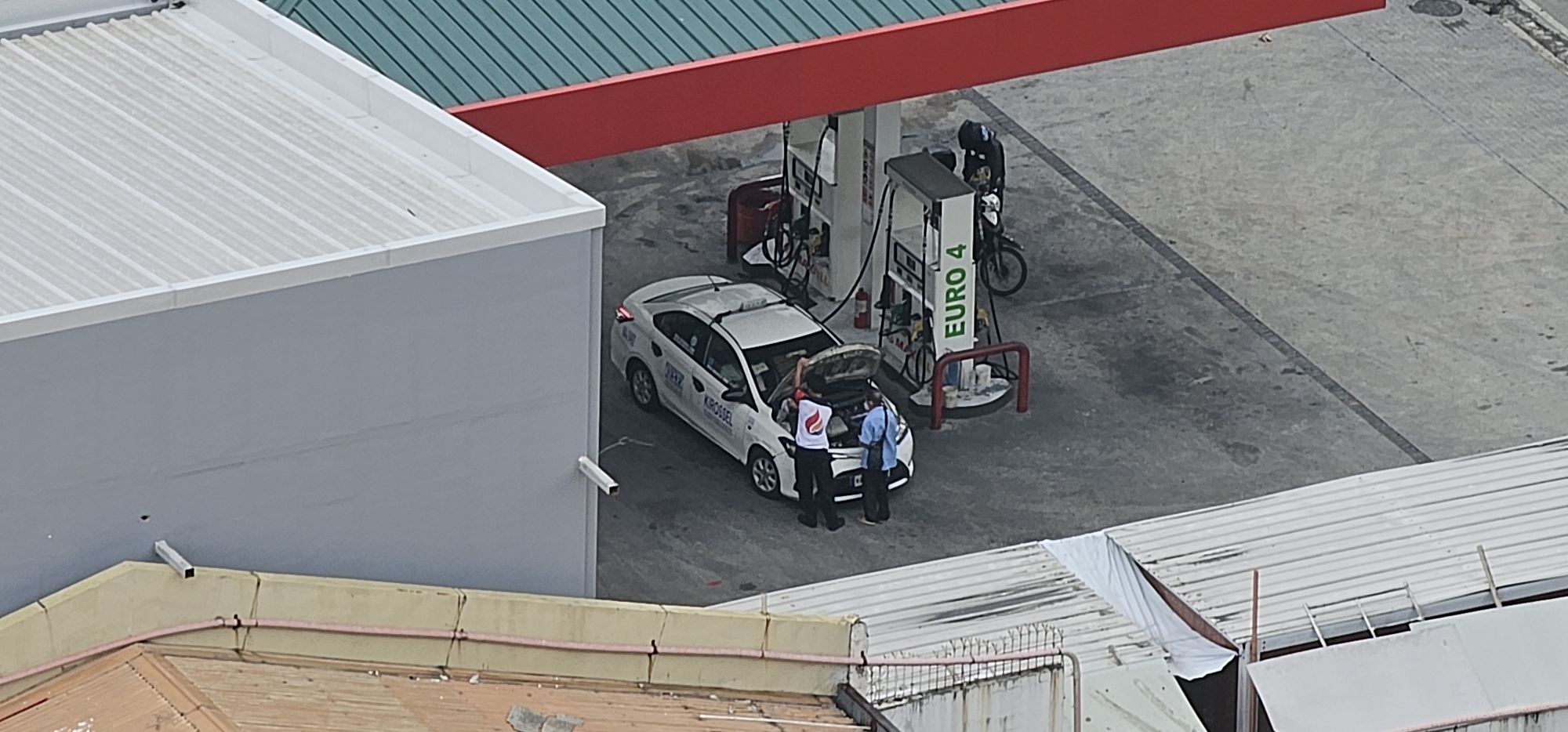An aerial view captures a gasoline station with a red canopy and green metal roofing, situated on a paved lot in Makati City, Philippines. Two individuals are interacting near the open hood of a white sedan parked beside a fuel pump labeled "EURO 4." One person, wearing a white shirt with a red and orange logo, appears to be gesturing towards the engine. Another person in a light blue shirt stands next to them, also looking at the car's engine. A motorcycle is parked near another fuel pump. The scene is set against a backdrop of urban structures, including the light gray facade of a building to the left and white corrugated roofs of adjacent structures in the foreground.