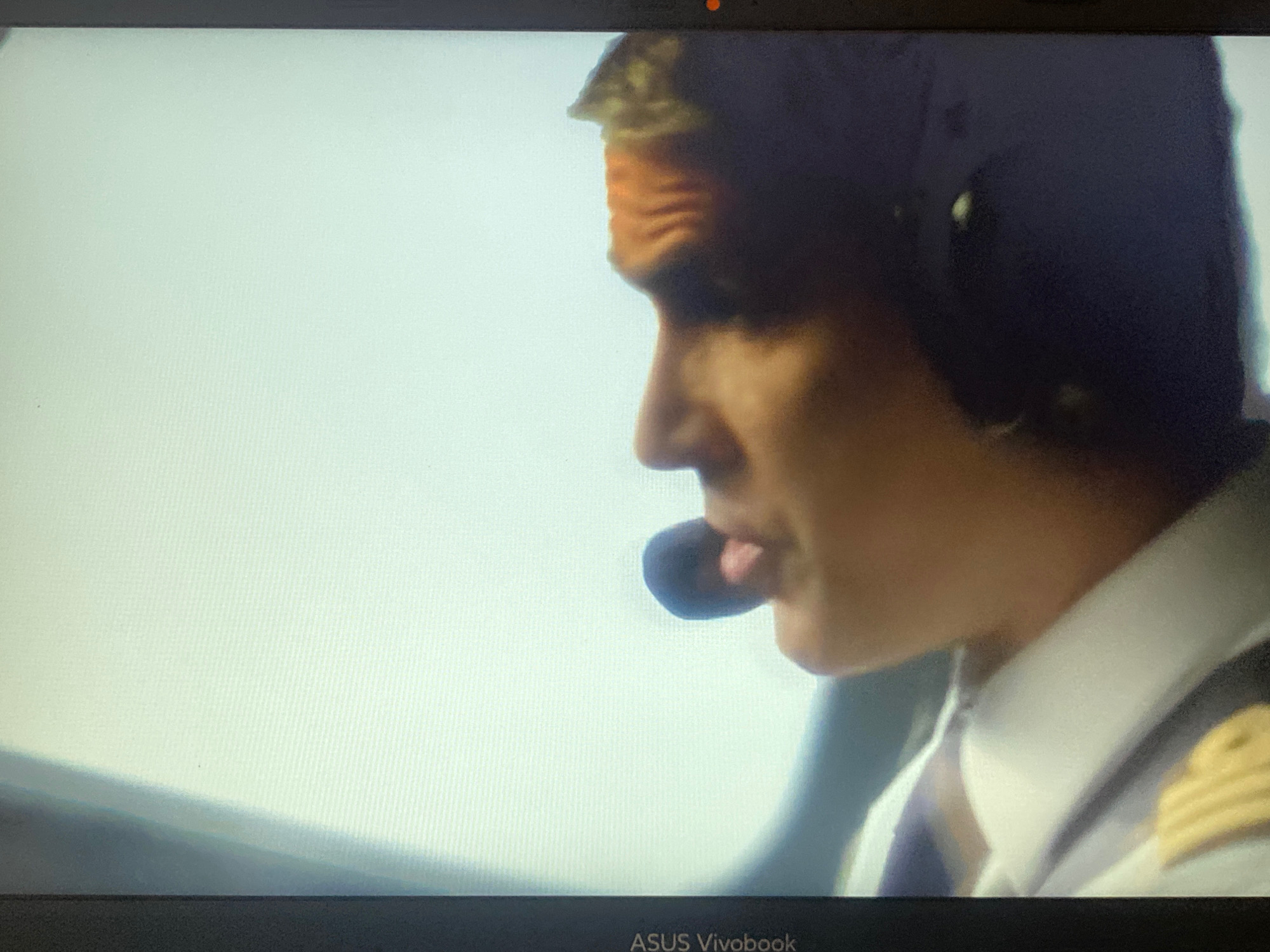 The image shows a close-up of a person wearing a pilot's uniform, including a headset with a microphone. The person's face is partially visible, with a determined expression. The background is a bright, featureless expanse, suggesting they are in the sky or a very bright environment. The person is looking to the left, with their mouth slightly open as if speaking. The pilot's uniform includes a white shirt with epaulets, indicating a rank. The image is a still from a screen, evidenced by the "ASUS Vivobook" text visible at the bottom. The provided crops offer no additional contextual information regarding the location in Hemayetpur, Bangladesh, or any specific details that can be inferred about the scene beyond what is visible in the primary image.