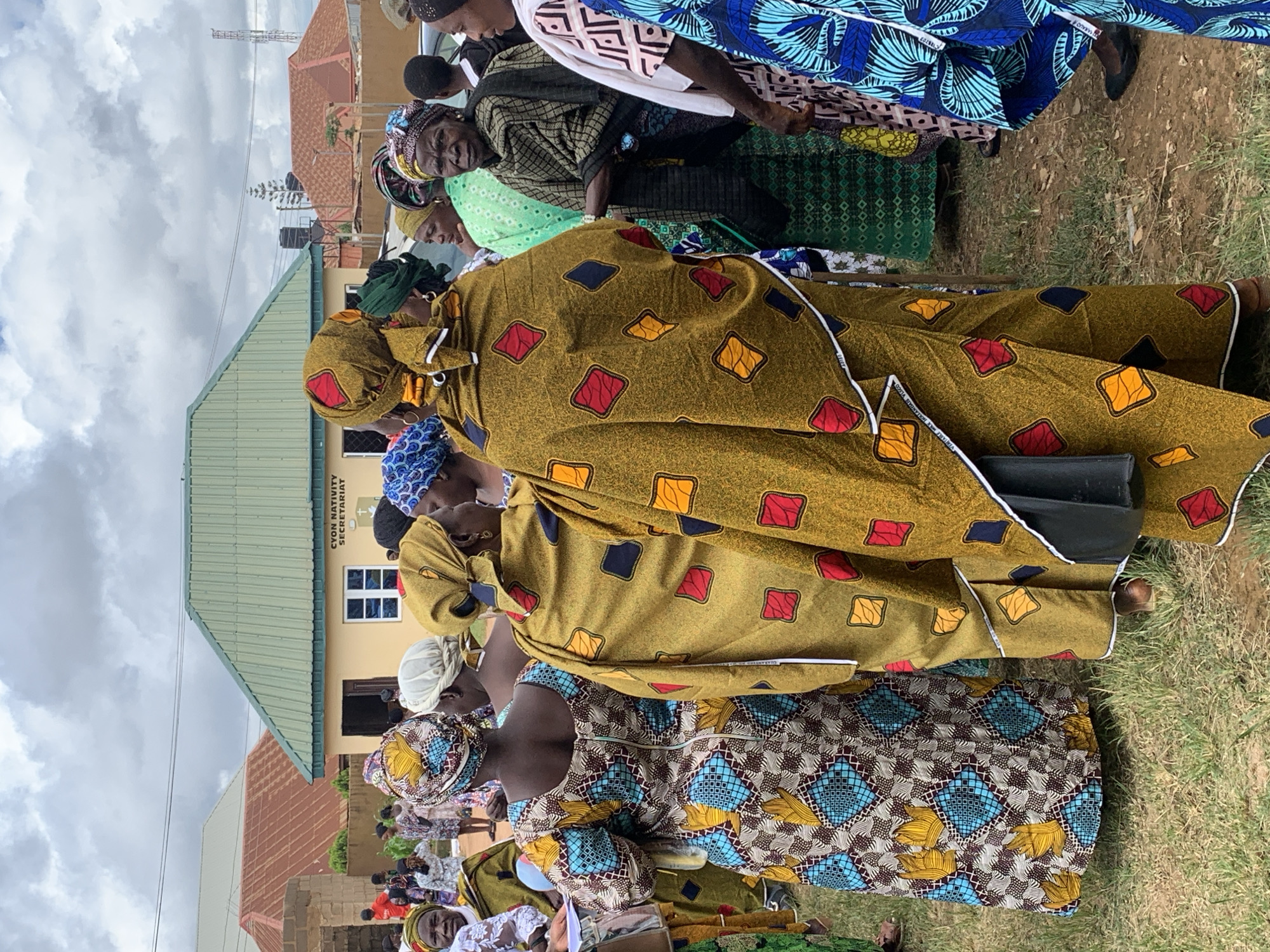 In Jos, Nigeria, a group of women, predominantly dressed in vibrantly colored traditional African attire, are gathered outdoors. The scene appears to be in front of a light-colored building with a green corrugated metal roof, marked with the inscription "CYON NATIVITY SECRETARIAT." Several women are facing away from the camera, showcasing the intricate patterns of their dresses and headscarves. One woman in the foreground wears a golden yellow outfit adorned with red, yellow, and blue diamond-shaped motifs. To her left, another woman is seen from the back, her dress featuring a complex geometric pattern of blue, yellow, and brown. The ground is covered with dry grass and some scattered dirt. In the background, more women are visible, along with glimpses of residential buildings with reddish-brown roofs and a distant telecommunications tower. The sky is overcast with clouds.
