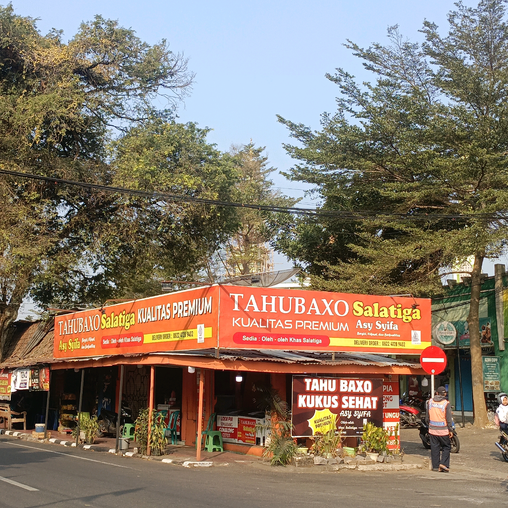 The image depicts a street scene in Salatiga, Indonesia, centered around a food stall named "Tahu Baxo Salatiga". The stall features a prominent orange banner with white lettering that reads "TAHUBAXO Salatiga KUALITAS PREMIUM" and "Asy Syifa". Below this, smaller text indicates "Sedia : Oleh - oleh Khas Salatiga" and a delivery order number "0822 4128 9402". Another banner to the right also advertises "TAHU BAXO KUKUS SEHAT" with "Asy Syifa" below it.
In the foreground, the street is paved with asphalt, and a sidewalk with a black and white striped pattern runs alongside. Various potted plants are placed in front of the stall. To the right of the stall, a red and white circular traffic sign prohibiting entry is visible. A person wearing an orange vest over dark clothing stands near the sign, facing away from the camera. Behind them, two individuals on motorcycles are also present, one of whom is a woman wearing a helmet.
The stall itself is a simple structure with a corrugated metal roof and open sides, revealing the interior where food items and seating appear to be arranged. Several other smaller signs and posters are attached to the facade of the stall.
In the background, tall trees with dense green foliage dominate the upper portion of the image, with clear blue sky visible between the branches. Power lines are strung between poles in the background. The overall atmosphere suggests a bright, sunny day.