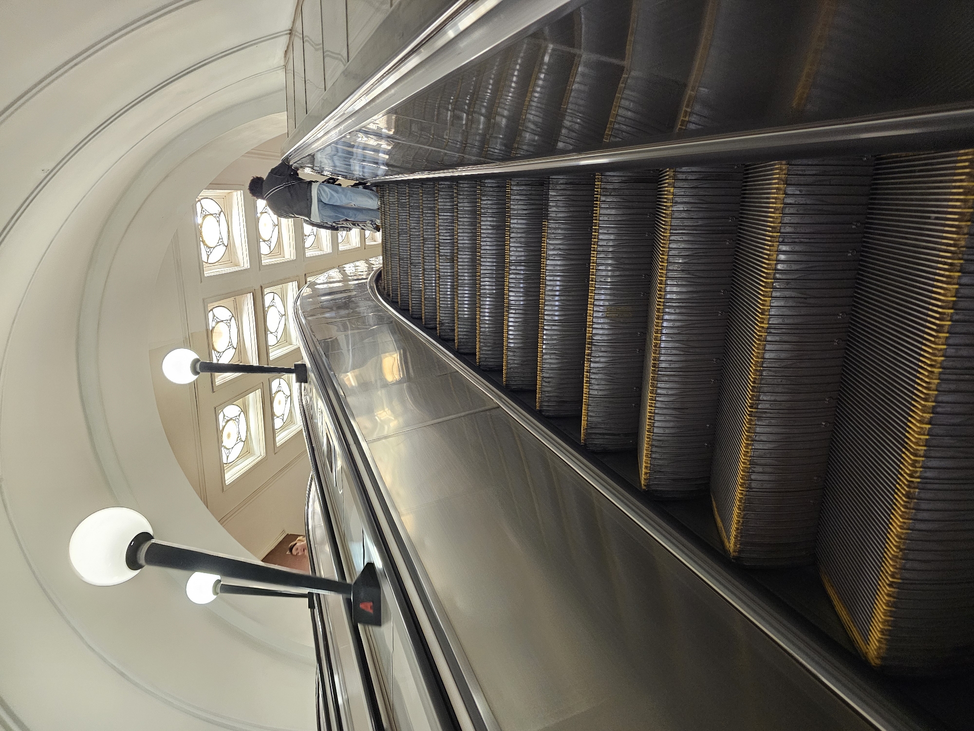 The image depicts an upward escalator ride at the Moscow Metro, specifically at the Komsomolskaya station. The escalator's structure consists of metallic steps and handrails, illuminated by globe-shaped lamps attached to black posts along the side. A person in a gray jacket and blue jeans is standing on the escalator near the top. The background is characterized by a rounded, white wall with square, stained-glass windows.