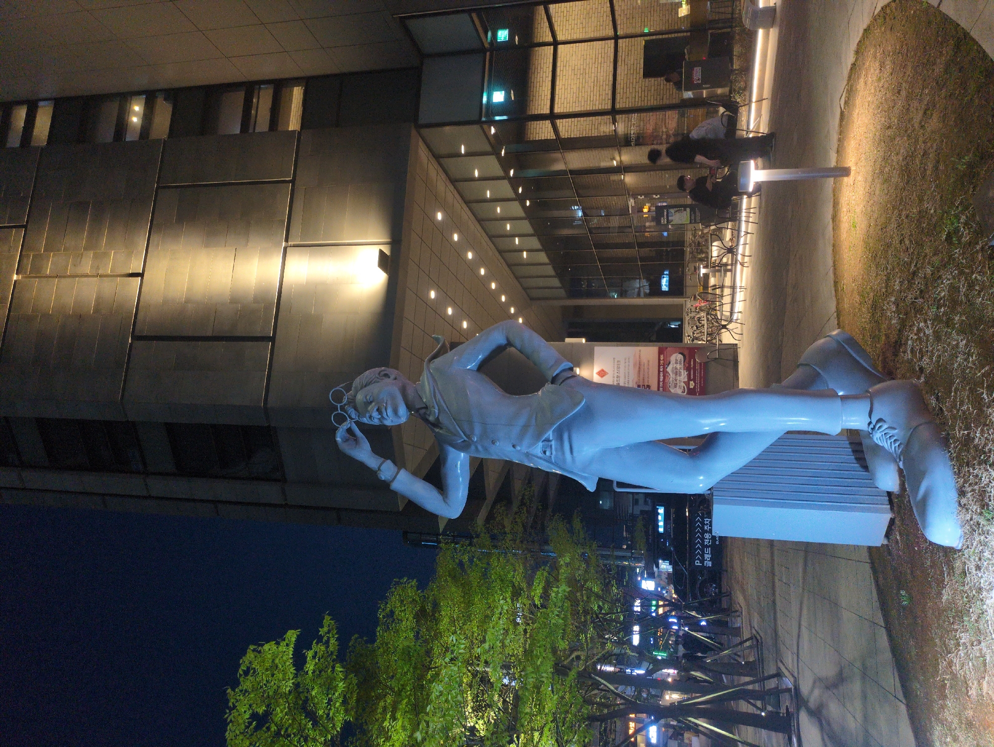 At night, a statue of a man is positioned in front of a modern building, potentially in Seoul, South Korea. The statue, a full-body depiction, is of a fair complexion. He's wearing a vest over a collared shirt, trousers, and shoes with laces. He holds a pair of round glasses near his face. He is posed lying on his side atop a gray rectangular structure. Behind the statue, there is a tree, its branches and leaves illuminated, likely by streetlights. In the background, there is the building, with its lit windows, and some outdoor seating with people. Part of the sidewalk and a patch of grass are visible around the statue's base. In the top left of the frame, the night sky transitions from a dark blue to almost black, indicating the late hour. A sign with Korean characters is partially visible beside the tree, reinforcing the possibility of the image's origin.