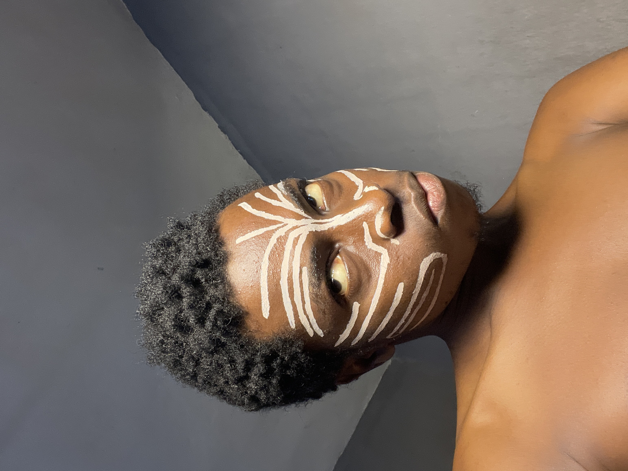 A close-up shot shows a person's face and upper body, with white face paint applied in a patterned design around the eyes and cheeks. The person has dark, curly hair and is looking directly at the camera with a neutral expression. The background is a plain, gray wall. This image was captured in Nkpor, Nigeria.