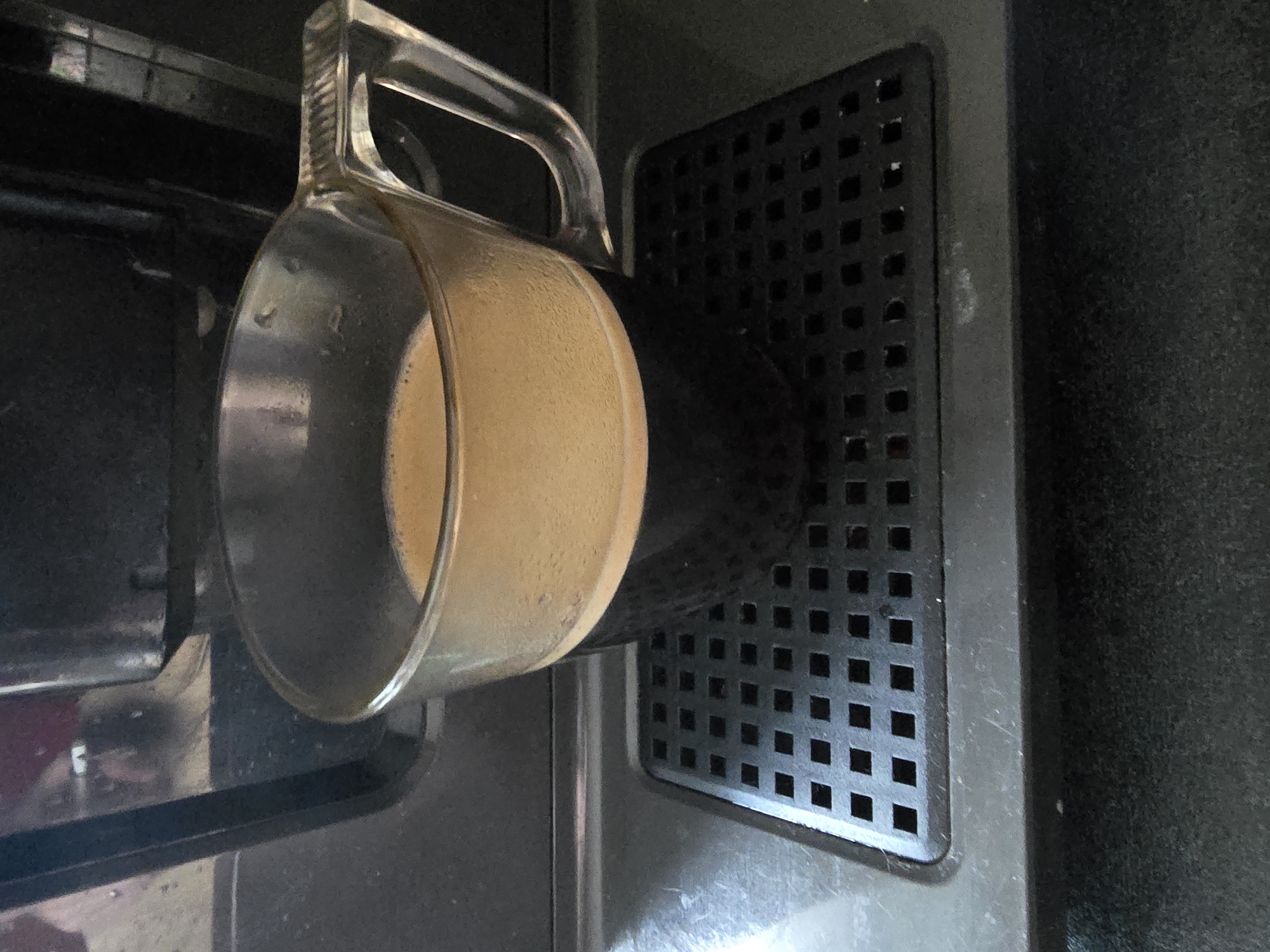 A clear glass cup filled with dark coffee and a foamy crema sits on a black, perforated drip tray. The coffee machine is visible in the background, with a portion of its dark exterior. The scene appears to be indoors, likely in a kitchen or coffee shop, given the presence of the coffee machine and the cup.