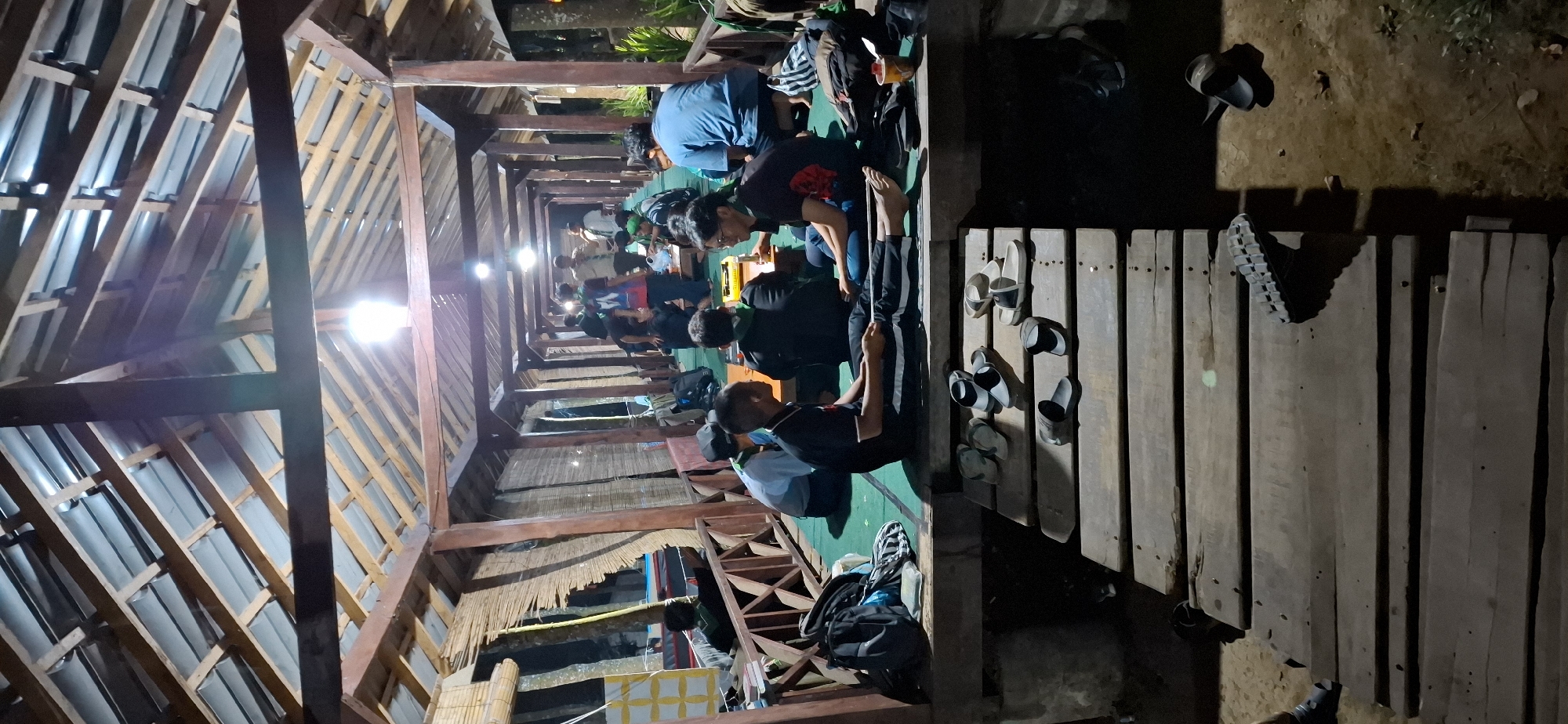 In Puntukrejo, Indonesia, a group of individuals are gathered in a covered, open-air structure with a wooden roof supported by pillars. The structure appears to be a pathway or a gathering space, with a wooden floor covered by a green mat. Several people are seated on the mat, some facing each other in conversation. One group in the foreground is engaged in what seems to be a discussion. Another group is visible further in the background, also interacting. There are several bags and backpacks scattered around the gathering area. On a lower wooden walkway in front of the main gathering space, numerous pairs of sandals are placed, suggesting that people have removed their footwear before entering the main area. The lighting in the scene is dim, with artificial lights illuminating the space from above. The environment suggests a relaxed, informal setting, possibly a communal area or a rest stop.