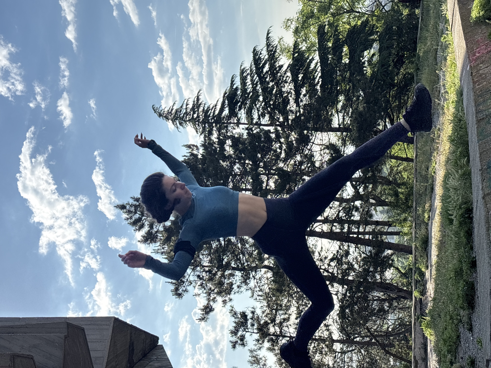 A person is captured in a dynamic, balancing pose outdoors in Varna, Bulgaria. The individual, dressed in a blue long-sleeve crop top and dark leggings, is performing an acrobatic or dance-like movement. They are balancing on one foot on a narrow concrete ledge, with their other leg extended high and arms outstretched.
The background features a bright blue sky dotted with scattered white clouds. Lush green trees, including tall conifers and some deciduous varieties, frame the scene, creating a natural setting. A concrete or stone structure, possibly a wall or railing, with grass and weeds growing alongside it, runs across the mid-ground and right side of the image, serving as the balance point for the person. Another geometric concrete structure is partially visible in the lower left corner.
The image appears to be taken during the daytime under clear weather conditions. The pose conveys a sense of strength, balance, and focused athleticism. No visible text is present in the image.