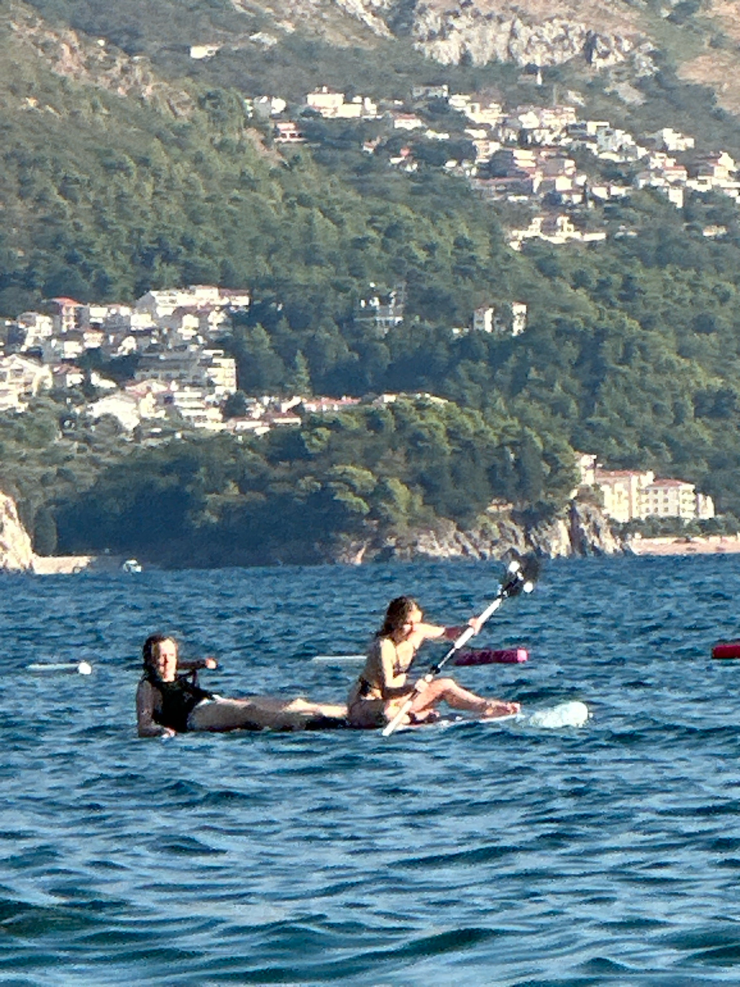 Two individuals are on a paddleboard in the Adriatic Sea, off the coast of Boreti, Montenegro. The person in the foreground is holding a paddle and appears to be paddling. The other person is lying down on the paddleboard. The water is blue with gentle waves. In the background, a coastal town with white buildings is visible on a hillside covered in green trees. The sky is clear and the sun is shining.