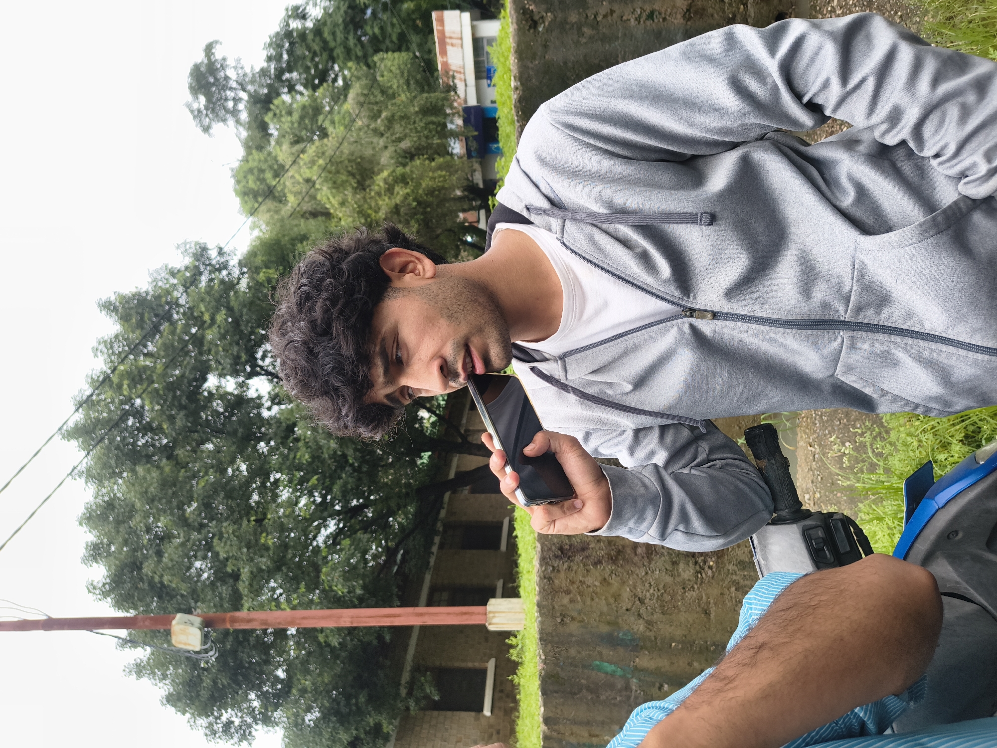 A young man with curly dark hair is holding a smartphone to his ear and speaking, with his mouth slightly open. He is wearing a gray hooded jacket over a white t-shirt. His left hand is resting on the handle of a blue scooter. Another person, wearing a blue and white striped shirt, is partially visible on the left, their arm resting on the scooter. They are standing outdoors in front of a low stone wall with grass and trees behind it. In the background, there is a building with a red-capped pole in front of it. The sky is overcast. This scene is located in Dharamshala, India.