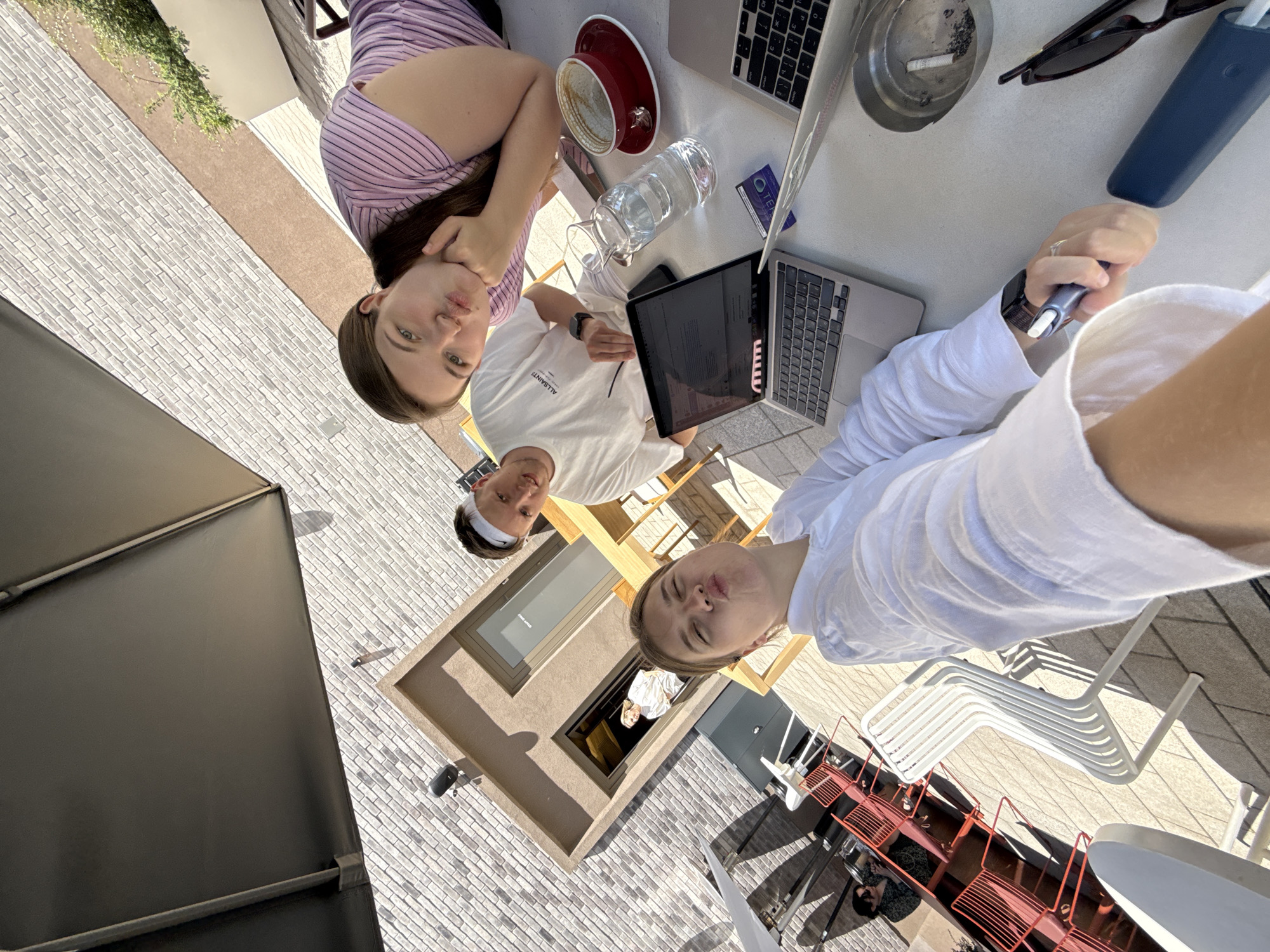 Three young adults are seated at a table on an outdoor patio, possibly a cafe or restaurant in Belgrade, Serbia. The patio is paved with light-colored bricks. The patio is adjacent to a building with a modern design, including a recessed window. An umbrella provides shade.
The table contains two laptops, a red coffee cup, a glass water bottle, an ashtray with a cigarette, and sunglasses. The first person, wearing a purple striped top, is in the upper left of the frame, leaning on her hand and looking at the camera. The second person, wearing a white "ALLSAINTS" t-shirt and a white headband, is situated near the center of the image, looking slightly to the left. The third person, wearing a white long-sleeved shirt, is holding a smoking device (possibly an IQOS) and is in the lower right part of the frame. Several white and red metal chairs are stacked nearby.
