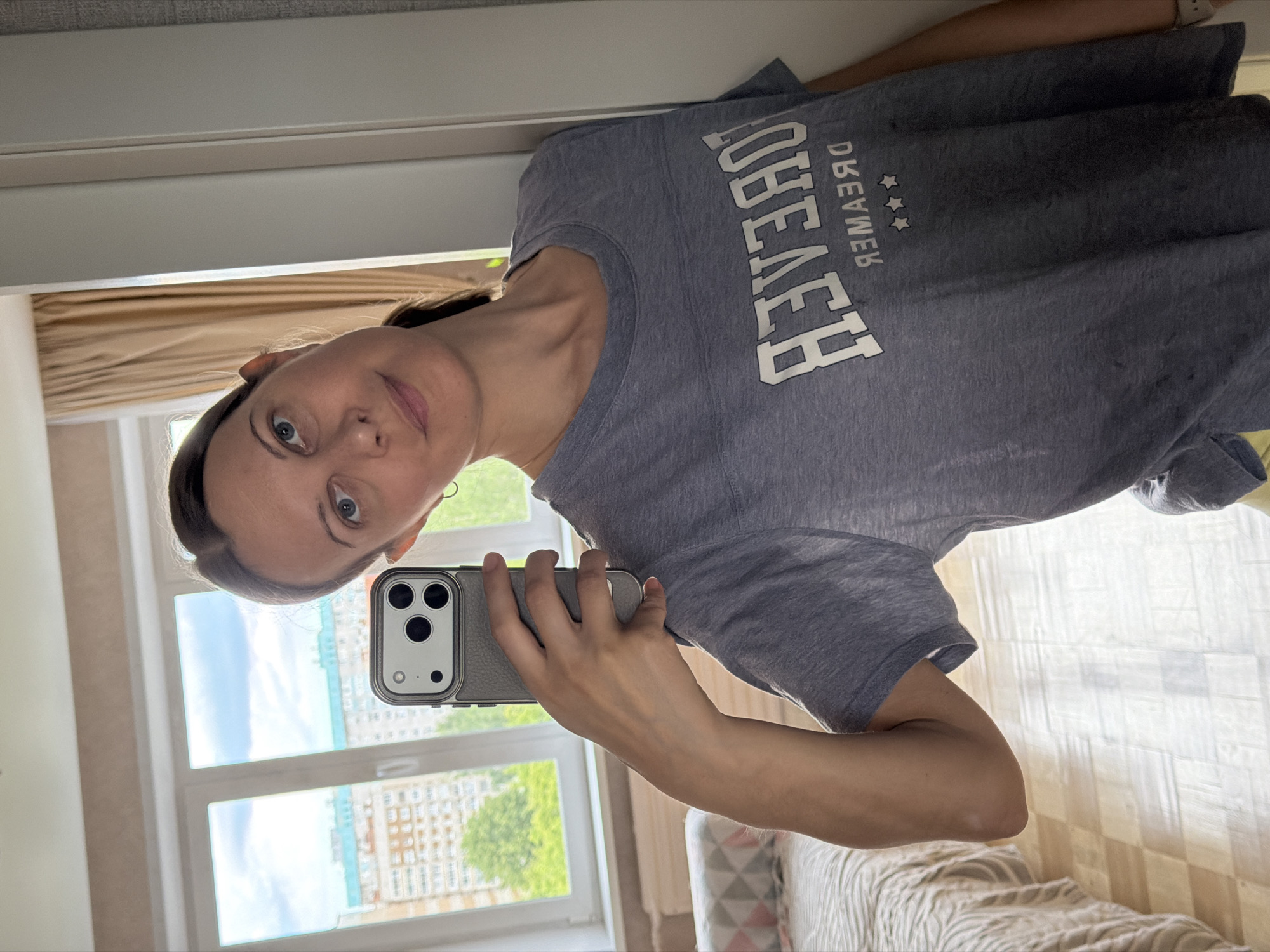 A woman is taking a selfie in a room that appears to be an apartment in Izhevsk, Russia. She is wearing a gray t-shirt with white lettering that reads "FOREVER DREAMER". The room has a bed with a patterned bedspread, a window with beige curtains showing a view of apartment buildings and trees, and a tiled floor. The woman is holding up a smartphone to take the picture.