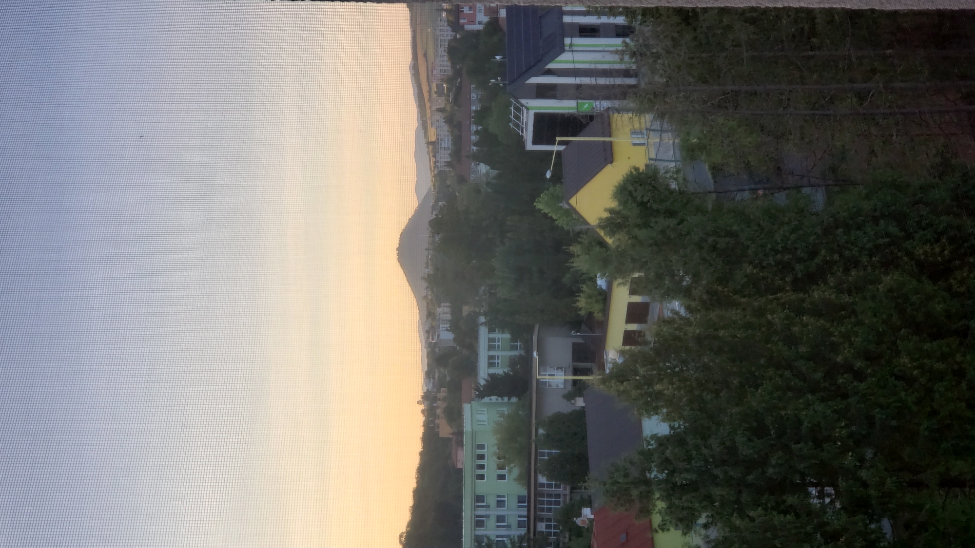 The image depicts a view of a city from a window, looking out at a hazy sunset. In the foreground, a mesh screen is visible, obscuring the view slightly. Beyond the screen, several trees with lush green foliage are prominent, with their branches and leaves creating a dense layer.
In the mid-ground, a variety of buildings are visible. There are multi-story residential buildings, some with light green facades and numerous windows. Other structures appear to be lower-rise commercial or institutional buildings, one of which is painted yellow with garages. A building with a dark roof and white and green vertical stripes is also present. Some smaller residential buildings with red roofs are partially visible.
In the background, a distinctive conical hill or mountain rises above the urban landscape. The sky exhibits a gradient of soft colors, transitioning from a pale blue at the top to a warm yellow and orange near the horizon, indicative of a sunset. Distant hills and more buildings are visible under the hazy sky. The overall impression is that of a suburban or urban fringe area at dusk. The image does not contain any people or animals.
This scene is likely located in Prešov, Slovakia, as the distinctive conical hill, known as the Prešovská