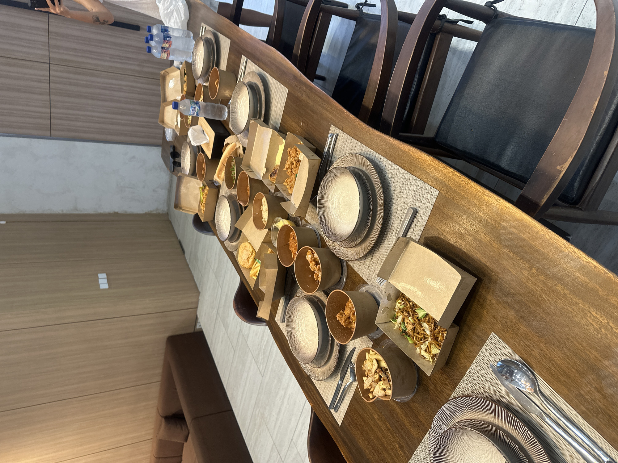 The image depicts a long wooden table set for a meal in what appears to be a modern dining area. The table is covered with individual placemats and settings for multiple people, including plates, cutlery, and brown paper containers holding food. The food seems to be a variety of takeout dishes. There are water bottles at one end of the table. Several dark brown dining chairs are arranged around the table. The background includes a wall with horizontal paneling. Based on the style of furniture and architecture, the scene may be located in Kuta Selatan, Indonesia, although this cannot be confirmed with certainty from the image alone. There is no visible human interaction. A sofa in the background indicates this may be a dining room in a private residence.