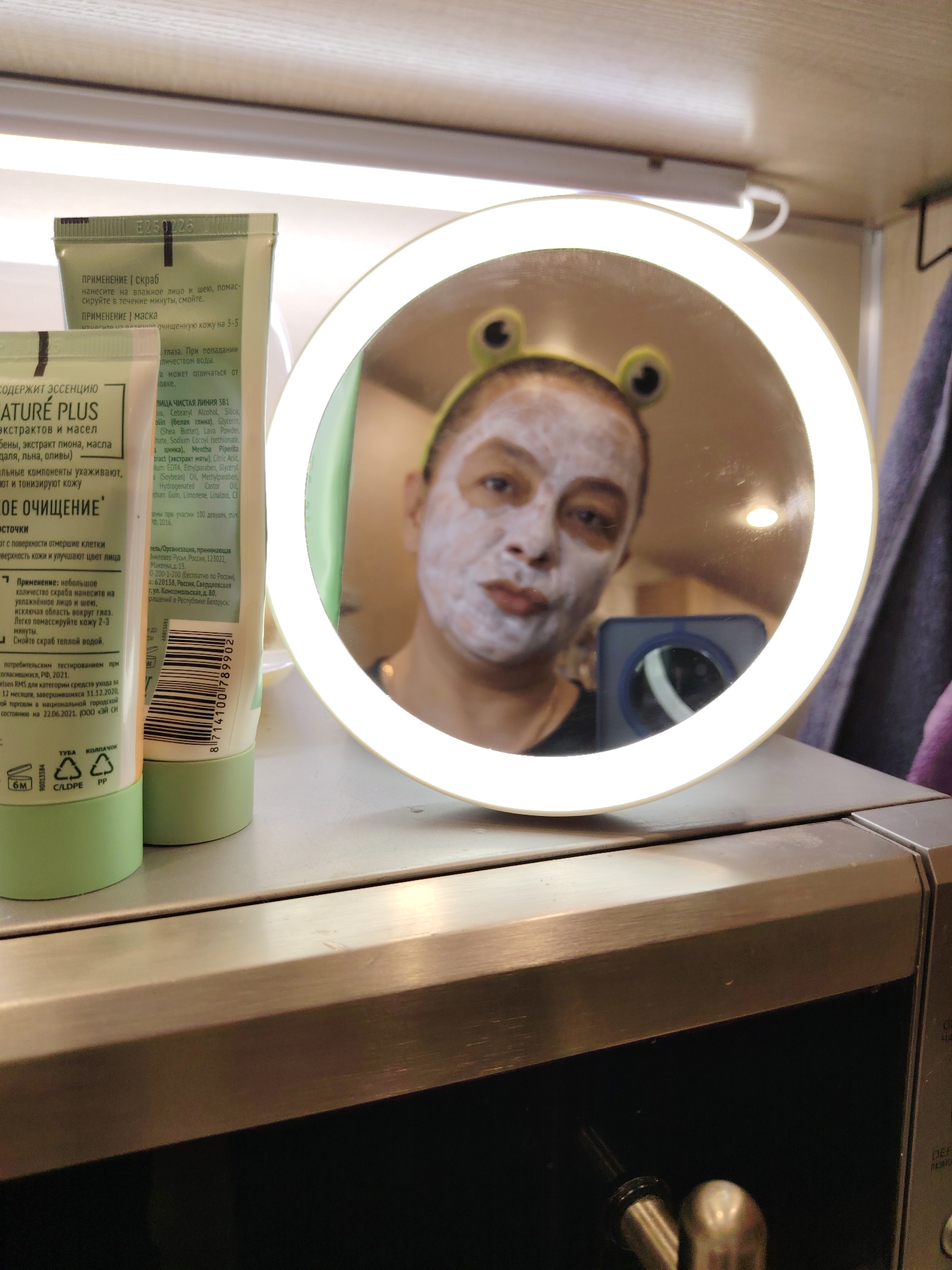 A close-up shot features a person reflected in a lighted makeup mirror, likely in a bathroom setting, as indicated by the presence of skincare tubes and a microwave oven. The person in the mirror is wearing a face mask and a headband with frog-like decorations. The mirror sits atop the microwave, with skincare tubes positioned to the left. The tubes feature Russian text and labels, specifically mentioning ingredients like "extracts of oils." The overall lighting is a mix of the mirror's integrated light and ambient lighting.