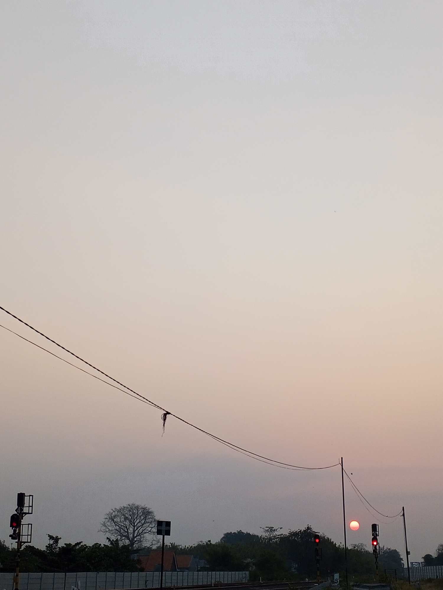 The image depicts a railway scene in Pakusamben, Indonesia, during what appears to be sunrise or sunset. The sky is a gradient of pale gray to light peach, suggesting a low sun position. Power lines stretch across the frame, with a piece of dark debris hanging from one of them.
In the foreground, a railway signal with a red light illuminated stands on the left. Further into the scene, another signal with a red light is visible, and to its right, the sun, a reddish-orange disc, is partially obscured by haze or a distant tree line. There are several trees, some bare and others with foliage, behind a low, grey wall or fence running along the railway tracks. A few buildings with red roofs are also visible amongst the trees. The overall impression is a quiet, possibly rural, industrial area at the beginning or end of the day.