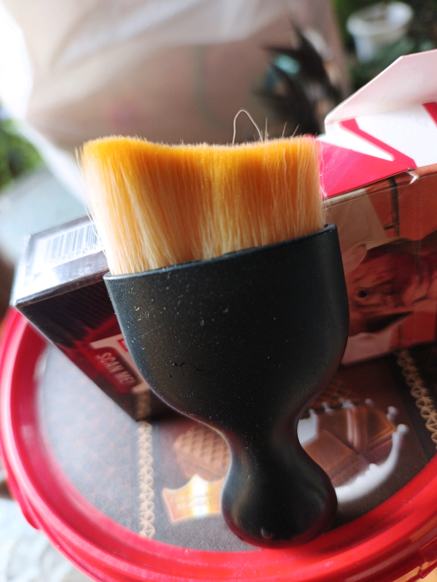 The image shows a close-up of a black-handled brush with orange bristles, resting on a red plastic rim. Behind the brush, partially visible, is a cardboard box with red and white markings, and another box with a barcode and the text "SCAN ME". The background is blurred, with hints of white fabric and greenery. The scene appears to be indoors, possibly on a table or counter, given the presence of packaging and the indoor lighting. The composition is focused on the brush, with the surrounding objects providing context. There are no people or animals visible in the image. The location is not specified as being in Ketegan, Indonesia.