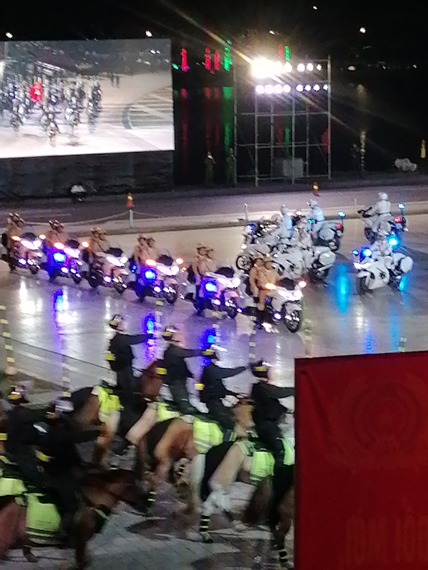 The image depicts an outdoor nighttime event in Da Lat, Vietnam, possibly a parade or demonstration. The scene features a large screen in the background displaying a black and white image of a crowd or procession. In the mid-ground, there are several police officers on motorcycles, their headlights and blue flashing lights illuminating the area. The motorcycles appear to be white, and the officers are wearing light-colored uniforms. In the foreground, there are several individuals on horseback, also appearing to be law enforcement, wearing dark uniforms and bright yellow vests. The ground surface is paved. The overall atmosphere is festive, with bright lights and a sense of organized activity.