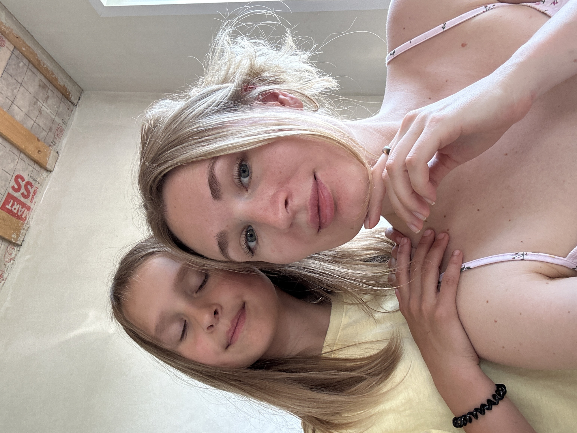 An overhead shot captures two females, one adult and one child, posing for a selfie indoors. The adult, positioned in the foreground, looks directly at the camera, her head tilted slightly, with her right hand touching her chin. The child is behind her, her eyes closed, with a serene expression and her left arm resting on the adult's shoulder. Both individuals have blonde hair. The background shows a wall and a ceiling with exposed wooden beams, suggesting ongoing construction or renovation. The image composition emphasizes the facial features of both subjects and their interaction.