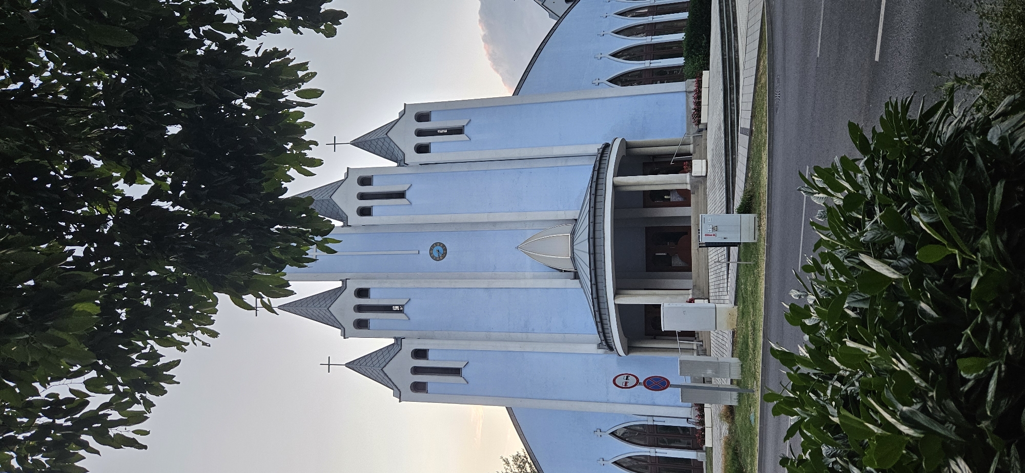 This is a vertical, eye-level, full shot of the Roman Catholic Church of the Holy Spirit in Hévíz, Hungary. The church has a distinctive light blue facade with white vertical accents and grey, pointed roofs on its towers. Two crosses are visible atop two of the towers. A clock face is centered on the middle tower. The church features arched windows and a circular portico with columns at the entrance. In the foreground, lush green bushes and a street are visible. Traffic signs are present on the left side of the image, including a no-entry sign and a no-parking sign. Electrical cabinets are also situated near the church entrance. The sky is overcast, suggesting it may be early morning or late evening. The image is taken from across a street, with foliage obscuring parts of the upper portion of the church.