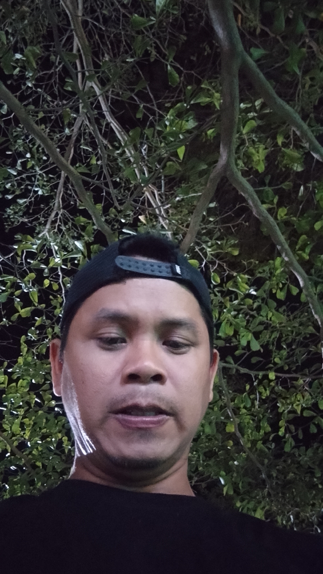 A close-up, low-angle selfie shows a man wearing a black baseball cap and a black t-shirt. He has brown skin and dark hair, and he is looking directly at the camera with a neutral expression. The background consists of tree branches and green leaves, suggesting an outdoor setting, possibly in a park or garden. The lighting is dim, with some artificial light illuminating the man's face and parts of the foliage. No specific landmarks or signs are visible to pinpoint the exact location within Karanglangu, Indonesia. The overall scene is casual and informal.