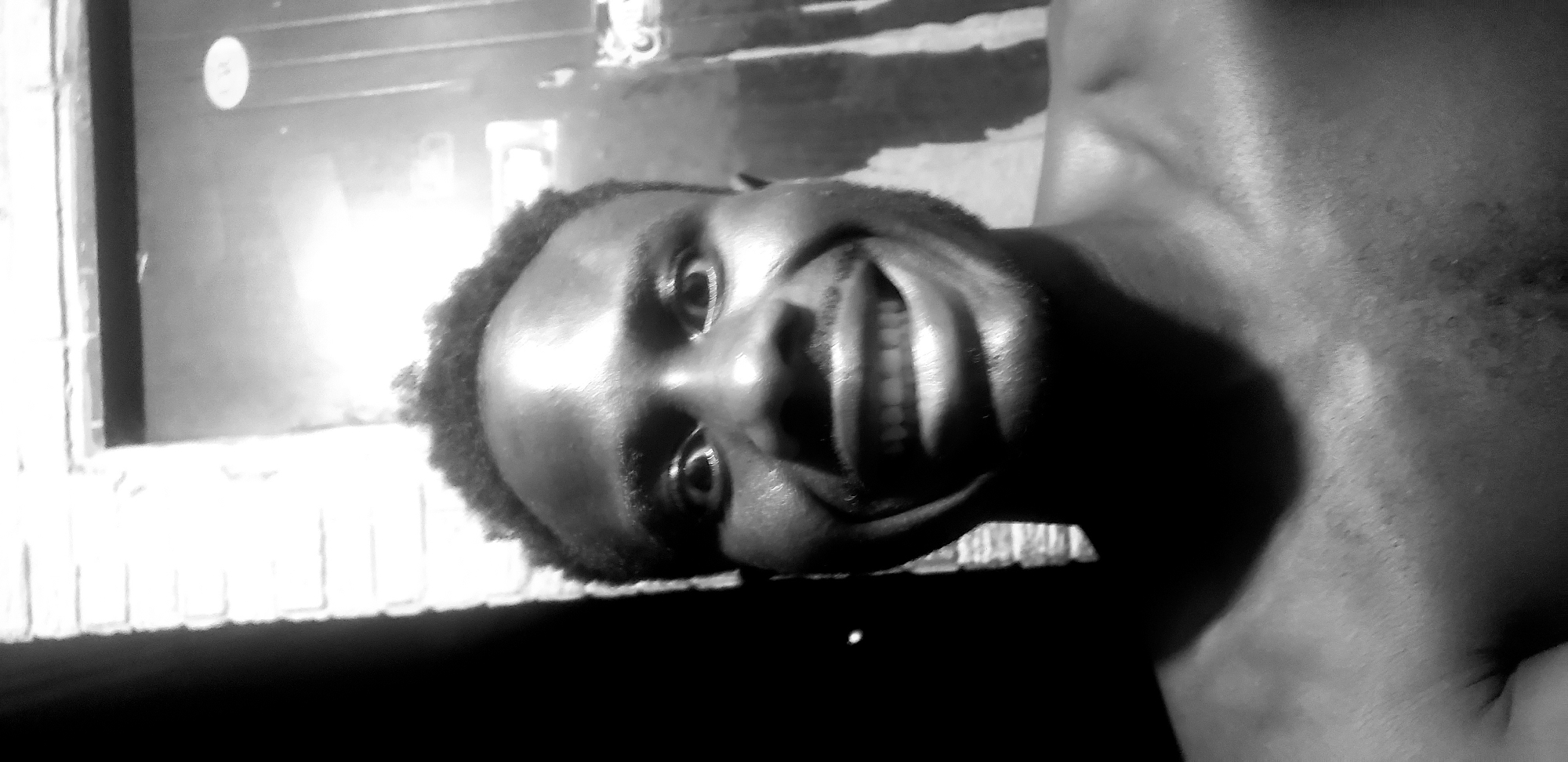 This is a black and white, close-up, sideways, selfie-style photograph of a person's face. The person has dark skin and curly hair. They are smiling broadly, showing their teeth. Their eyes are open, and they are looking towards the camera. The lighting is harsh, creating strong highlights and shadows on their face.
The background is out of focus and appears to be a window or a reflective surface. There are some indistinct shapes and patterns visible in the background, but they are not clear enough to identify specific objects. The overall impression is of a spontaneous and possibly playful moment captured by the person themselves.
There are no discernible text elements in the image. The setting appears to be indoors, given the lighting and the reflective surface in the background. The time of day and weather conditions are not evident. The emotion conveyed is one of joy or amusement.