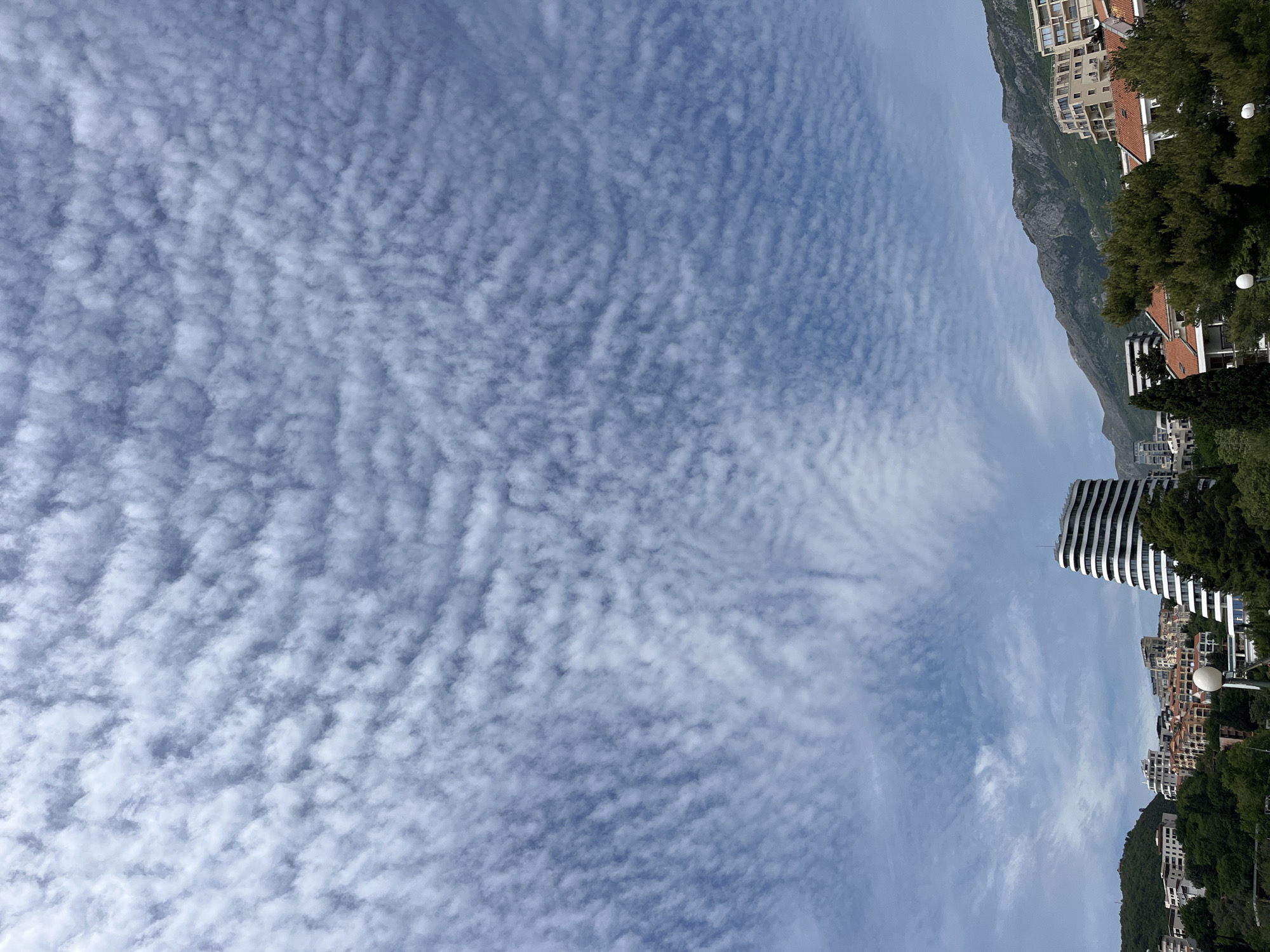 The image depicts a sky full of altocumulus clouds above the city of Boreti, Montenegro. In the background, a mountain range rises behind the city's buildings. A modern, striped building stands out among the more traditional architecture. Street lamps are visible along the city's edge, surrounded by greenery. The sky is primarily light blue, interspersed with the textured pattern of the clouds.