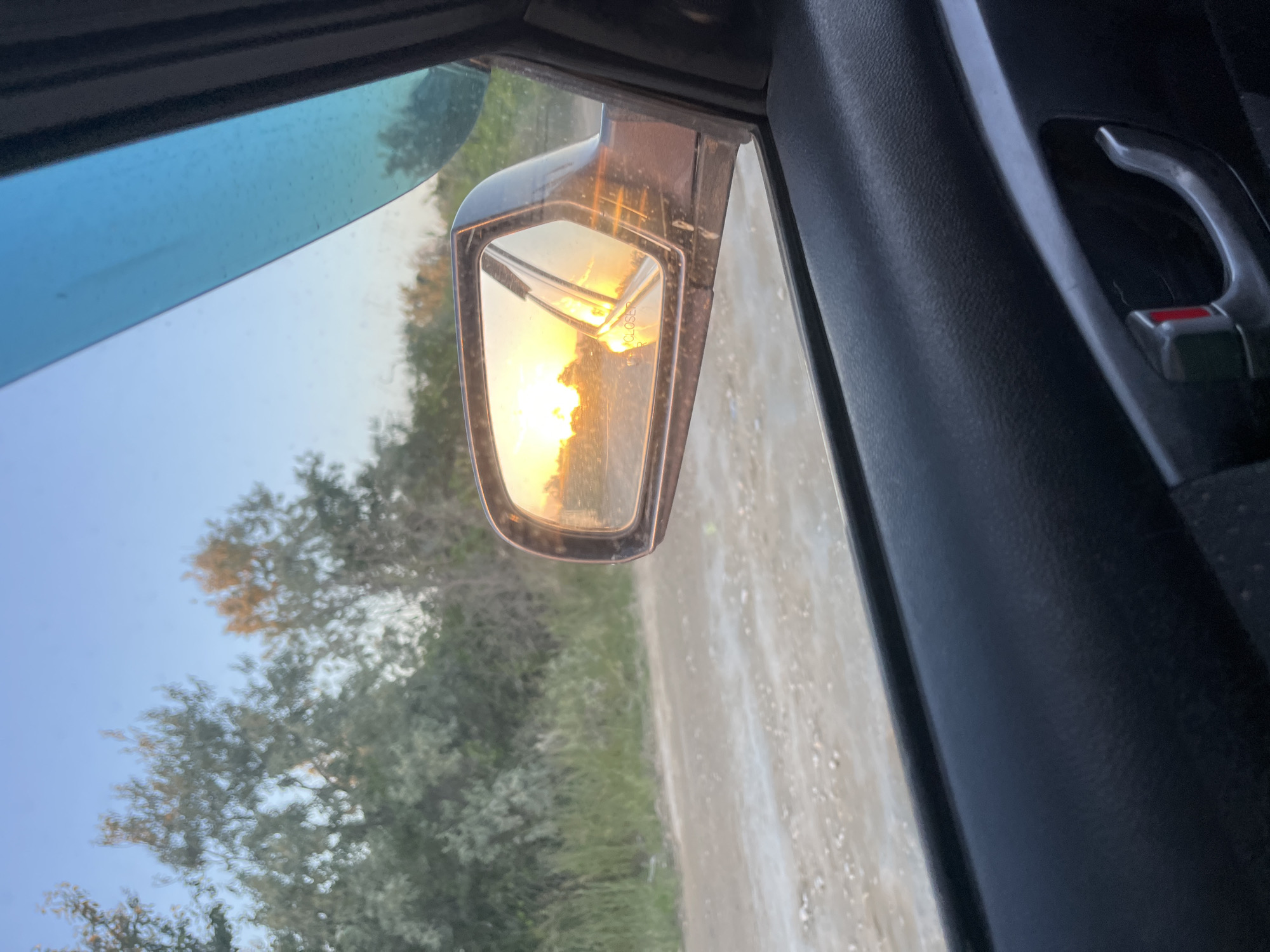 The image shows the view from inside a vehicle, focusing on the side mirror. The mirror reflects a scene of a dirt road or path running alongside foliage. The lighting in the mirror suggests either sunrise or sunset. A tree and the side mirror are clearly visible. The vehicle's interior, including the door panel and handle, are partially visible. The location cannot be confirmed as Kaspiysk, Russia without further information.