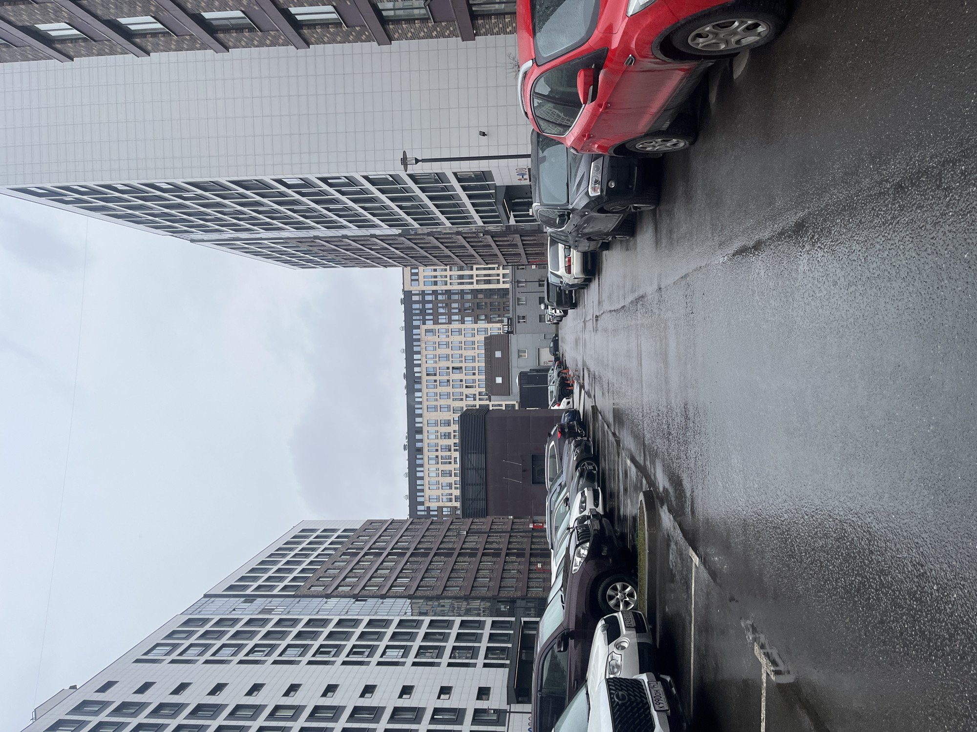The image depicts a rainy street in Saint Petersburg, Russia, lined with parked cars and tall buildings. The road surface is wet and reflects the overcast sky. The architecture is modern, with buildings featuring geometric patterns and neutral colors. A mix of vehicles is parked along both sides of the street. The scene appears to be an urban residential area.