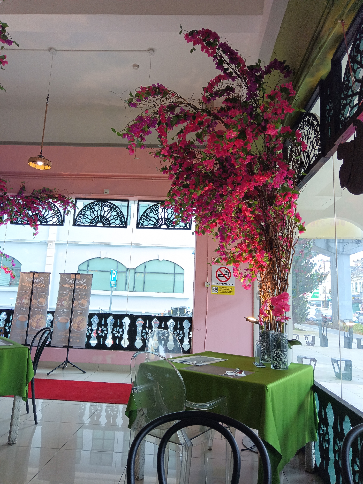 The image depicts the interior of a restaurant in Kuala Lumpur, Malaysia, with a focus on a decorative artificial tree adorned with vibrant pink and purple bougainvillea flowers. The restaurant features a modern aesthetic with transparent chairs and tables draped in green tablecloths. Large windows offer a view of the cityscape, including buildings and parked cars, suggesting an urban setting. On the wall, a "No Smoking" sign is visible, indicating a public establishment. The lighting appears to be a mix of natural light coming through the windows and artificial lights, including a pendant lamp hanging from the ceiling. The floor is tiled and reflects the surrounding elements.