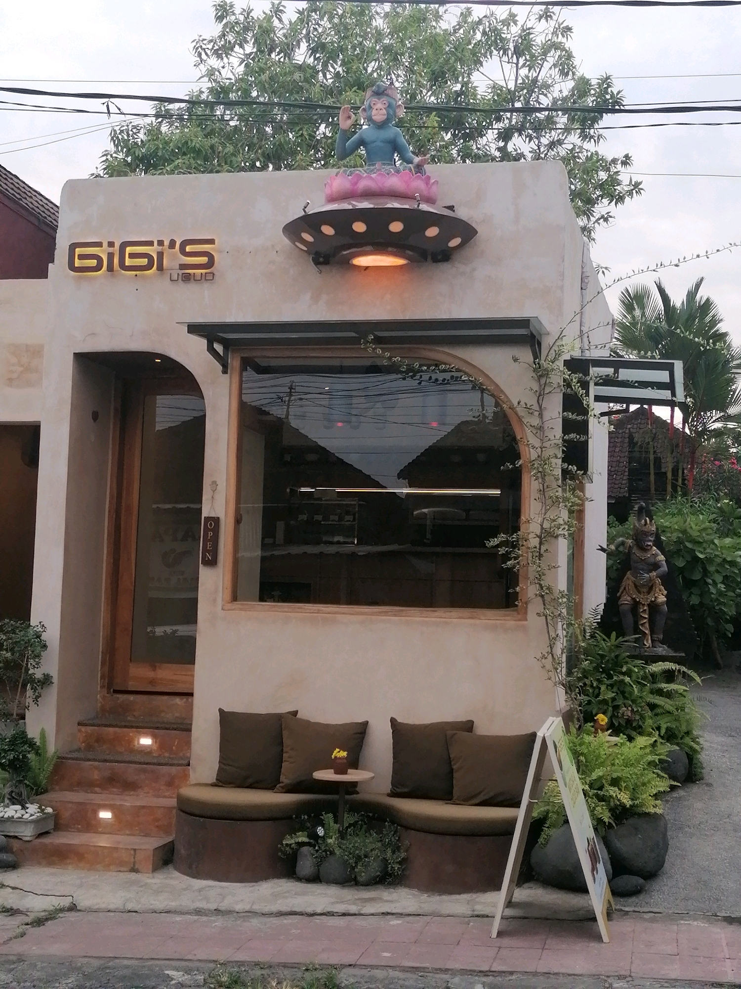 The image shows the exterior of a cafe or restaurant named "GiGi's Ubud" located in Ubud District, Indonesia. The building has a light-colored stucco facade with a wooden door and a large arched window. Above the entrance, there is a neon sign that spells out "GiGi's" in yellow. On the roof, a decorative statue of a monkey meditating on a lotus flower, which is placed on a saucer-like structure with lights, is visible. To the right of the entrance, there is a U-shaped seating area with brown cushions and two pillows. A small round table with a yellow flower in a vase sits on the seating. In front of the seating area, there is an A-frame sign with colorful graphics. To the right of the building, a statue of a Balinese figure in a golden costume is placed amongst lush green plants. Steps leading up to the wooden door are illuminated by small square lights. The environment appears to be an outdoor setting with vegetation and paved ground.