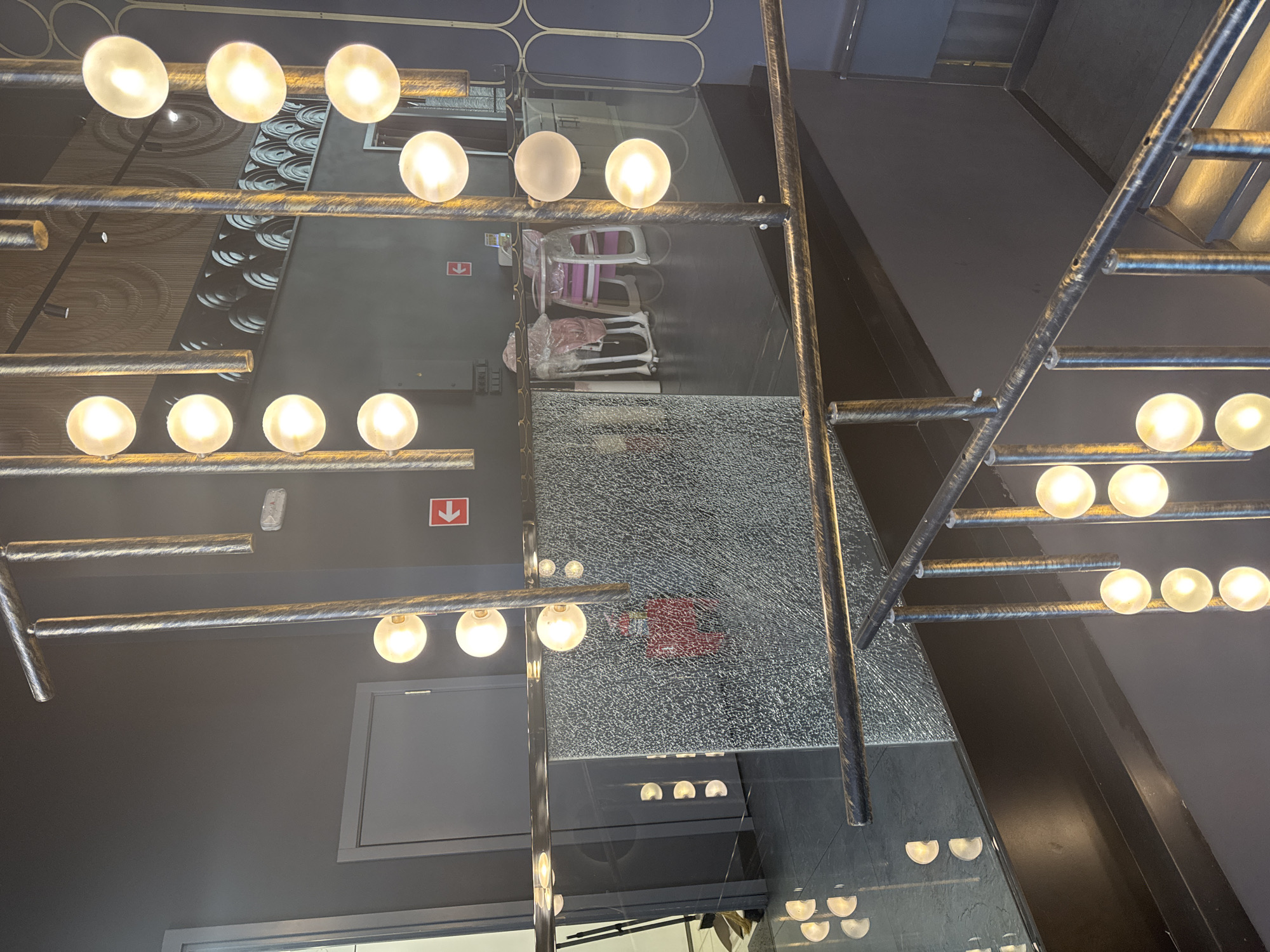 The image shows an interior view of a dimly lit, modern establishment, possibly a restaurant or lounge, in Moscow, Russia. A prominent feature is a multi-tiered chandelier with numerous glass orb lights, suspended from a textured, metallic ceiling. The chandelier's supports are metallic poles with a distressed finish. A reflective surface, likely a mirror or polished material, is positioned vertically in the foreground, creating a fragmented view of the scene.
The background reveals a dark gray wall with a door on the left and a red directional arrow sign pointing left. Further back, a row of what appear to be wine barrels or decorative circular elements are visible on a shelf, suggesting a wine cellar or tasting area. In the middle ground, there are two high chairs, one pink and white and the other white, covered in plastic wrap. Beyond these chairs, a counter area with kitchen equipment is partially visible.
A railing with a similar distressed metallic finish to the chandelier runs through the foreground, suggesting a staircase or mezzanine. The floor in the foreground is dark and reflective, mirroring the lights from the chandelier. The overall ambiance is one of contemporary design with a focus on industrial and metallic elements.