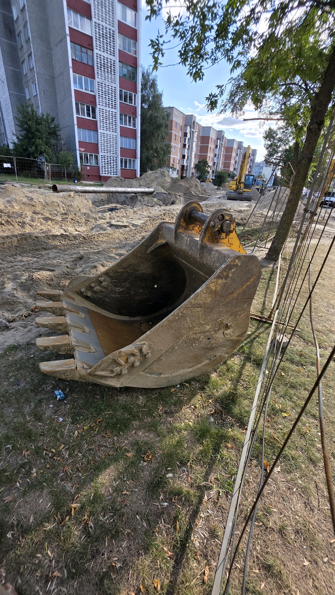 The image depicts a construction site with large building complexes in the background. In the foreground, a massive excavator bucket lies on the ground. The bucket is made of heavy-duty metal, showing signs of wear and tear. It has several sharp teeth along its front edge. The ground around the bucket is a mixture of sand and dry grass, with some fallen leaves scattered about. Behind the bucket, a trench with a large pipe running through it is visible. Further in the background, a yellow excavator is parked, and several multi-story residential buildings are lined up, suggesting an urban or suburban setting. A tall tree with green leaves stands to the right of the image, partially obscuring the view of the buildings. A wire fence runs along the right side of the frame, separating the construction site from the grassy area. The sky above is partly cloudy. The scene appears to be captured during daylight hours.