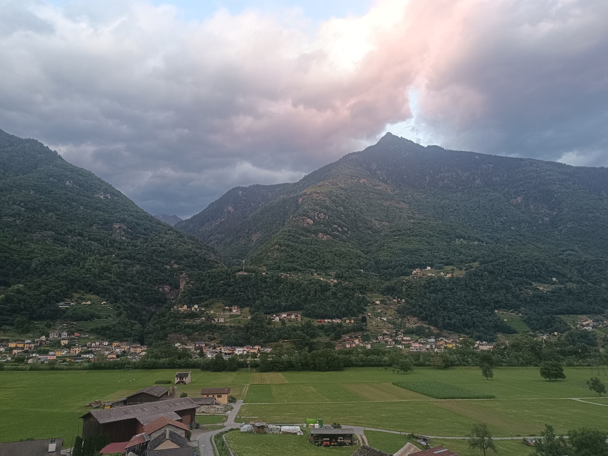 The image depicts a valley landscape in Serravalle, Switzerland, with a prominent mountain range dominating the background. The sky above is filled with dramatic, dark, and light grey clouds, with some areas of pinkish-white light suggesting either sunrise or sunset. The mountains are densely covered with lush green trees, with some scattered buildings visible on their slopes. In the foreground, there is a wide expanse of green fields, some appearing to be cultivated with distinct rectangular plots. Several farm buildings with pitched roofs are scattered across the fields, along with a winding road. The overall scene suggests a rural and mountainous environment under a cloudy sky.