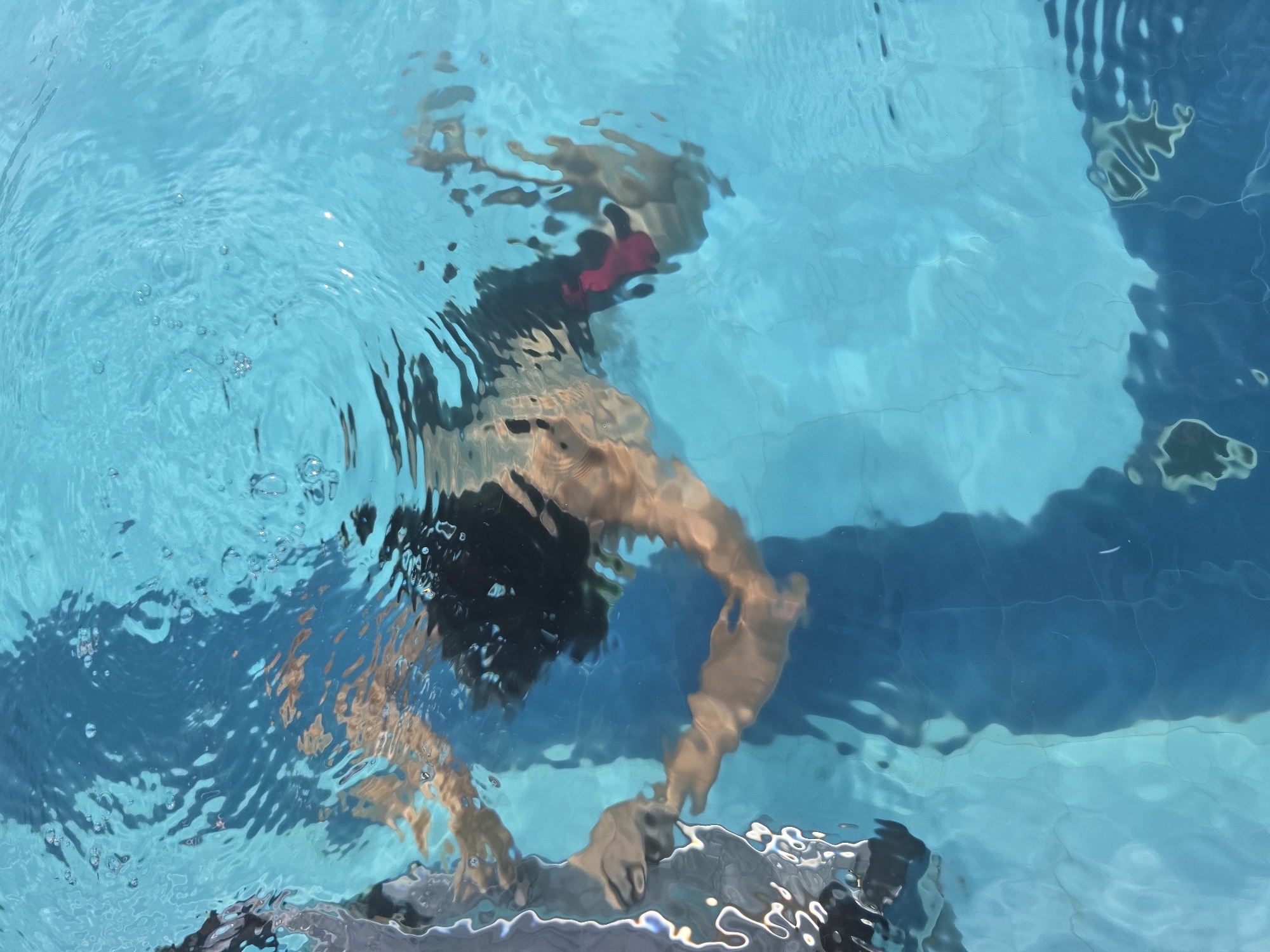 A person is swimming underwater in a swimming pool in Phu Thuy Ward, Vietnam. The water distorts the person's features and creates a rippling effect. The person is wearing a black swimsuit with a red accent. The pool's bottom is visible with different shades of blue.
