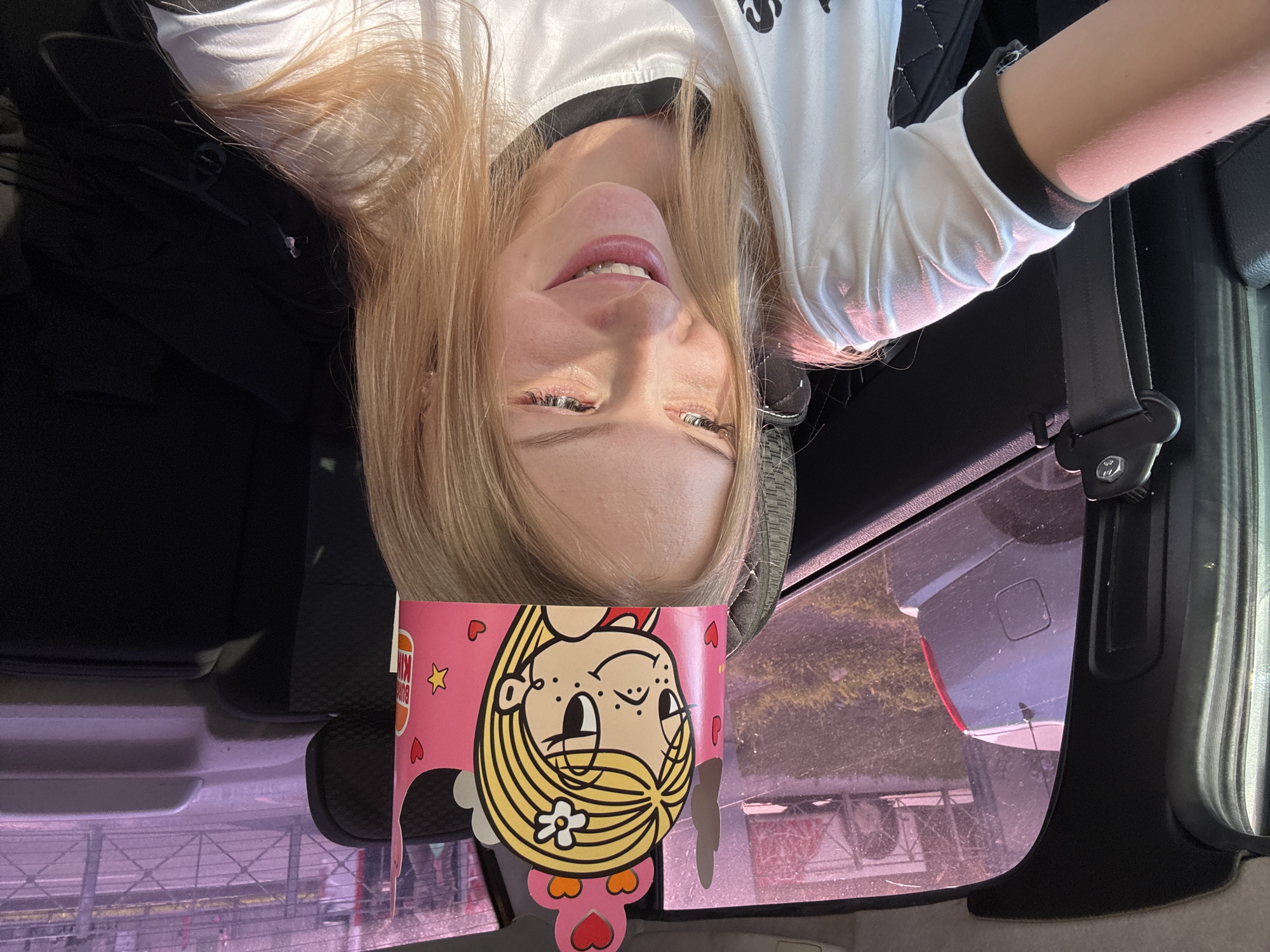 A young woman with blonde hair is wearing a pink paper crown with a cartoon character on it. She is sitting in the back seat of a car, with the passenger seat in front of her visible on the left side of the image. The woman is looking down and to the right, with a smile on her face. Her white t-shirt has black trim around the collar and sleeves. The sun is shining through the car window, casting a warm glow on her face. Outside the car window, a silver car is parked on a grassy area next to a fence. The scene appears to be outdoors, possibly in a parking lot or residential area. The specific location in Perm, Russia is not discernible from the image.