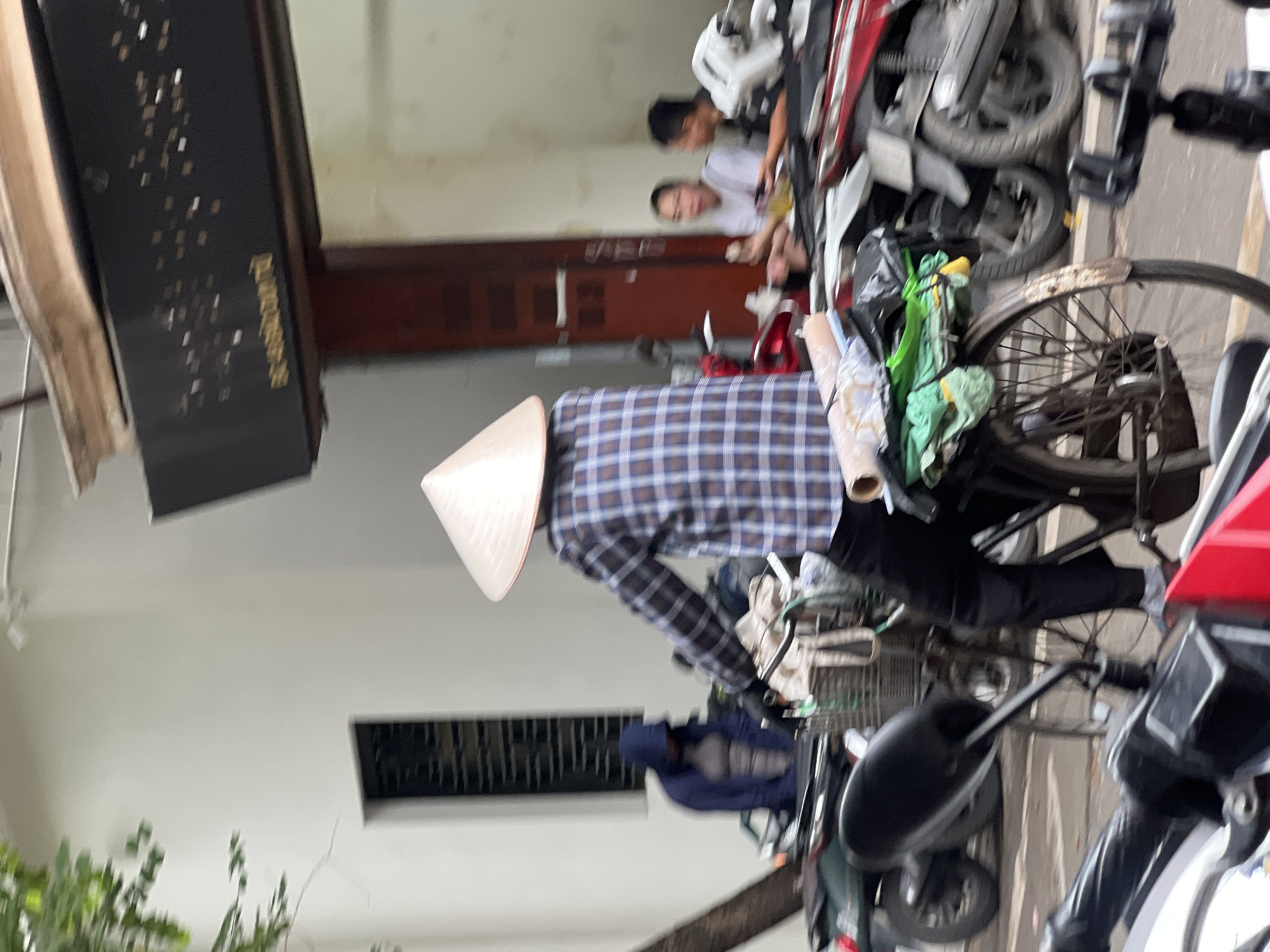 The image depicts a street scene in Hanoi, Vietnam, with a person wearing a conical hat riding a bicycle laden with items. The cyclist is dressed in a blue and white plaid shirt and black pants. Behind the cyclist, several motorcycles are parked. Further in the background, two individuals are seated, seemingly engaged in conversation or eating, with one holding a bowl. A building with a dark facade and decorative elements is visible above the seated individuals. The overall environment suggests a typical urban setting in Hanoi, likely a street or a pathway adjacent to businesses.