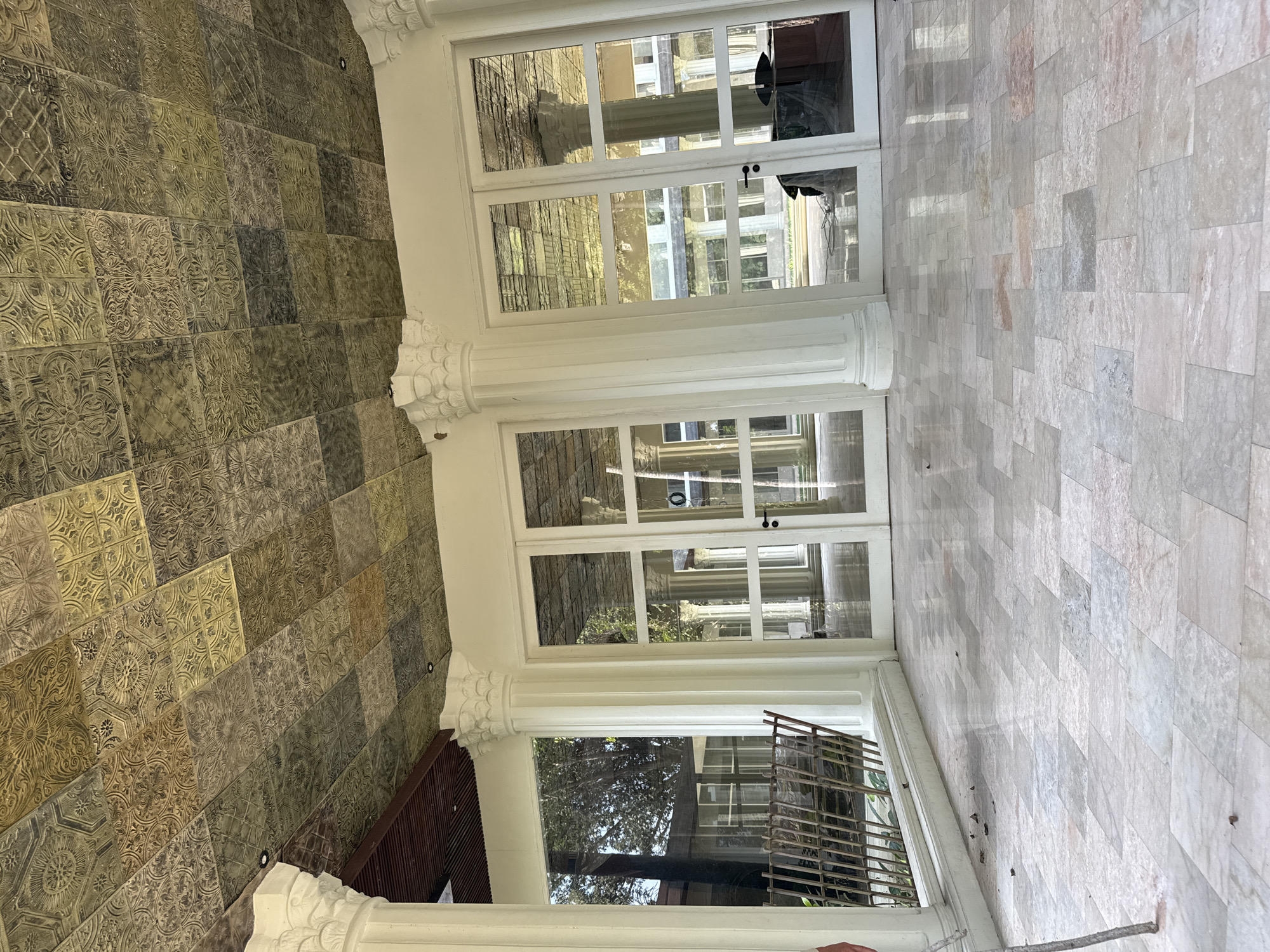 The image depicts an outdoor patio or covered walkway with a marble tile floor and a decorative metal ceiling. Large white pillars with ornate capitals support the structure. To the left, a metal fence is visible, with foliage and trees seen through a large glass window. In the center and right of the image, there are several glass doors leading into an interior space. Reflections in the glass doors show more pillars and architectural elements, suggesting a larger building or complex. The ceiling is composed of square tiles with intricate embossed patterns, varying in color from golden yellow to dark green and brown, giving it a somewhat aged appearance. The overall setting appears to be a well-maintained, possibly historical or luxurious, outdoor architectural space. The specific location of Tampaksiring, Indonesia, is not directly evident from the visual information in the image itself.