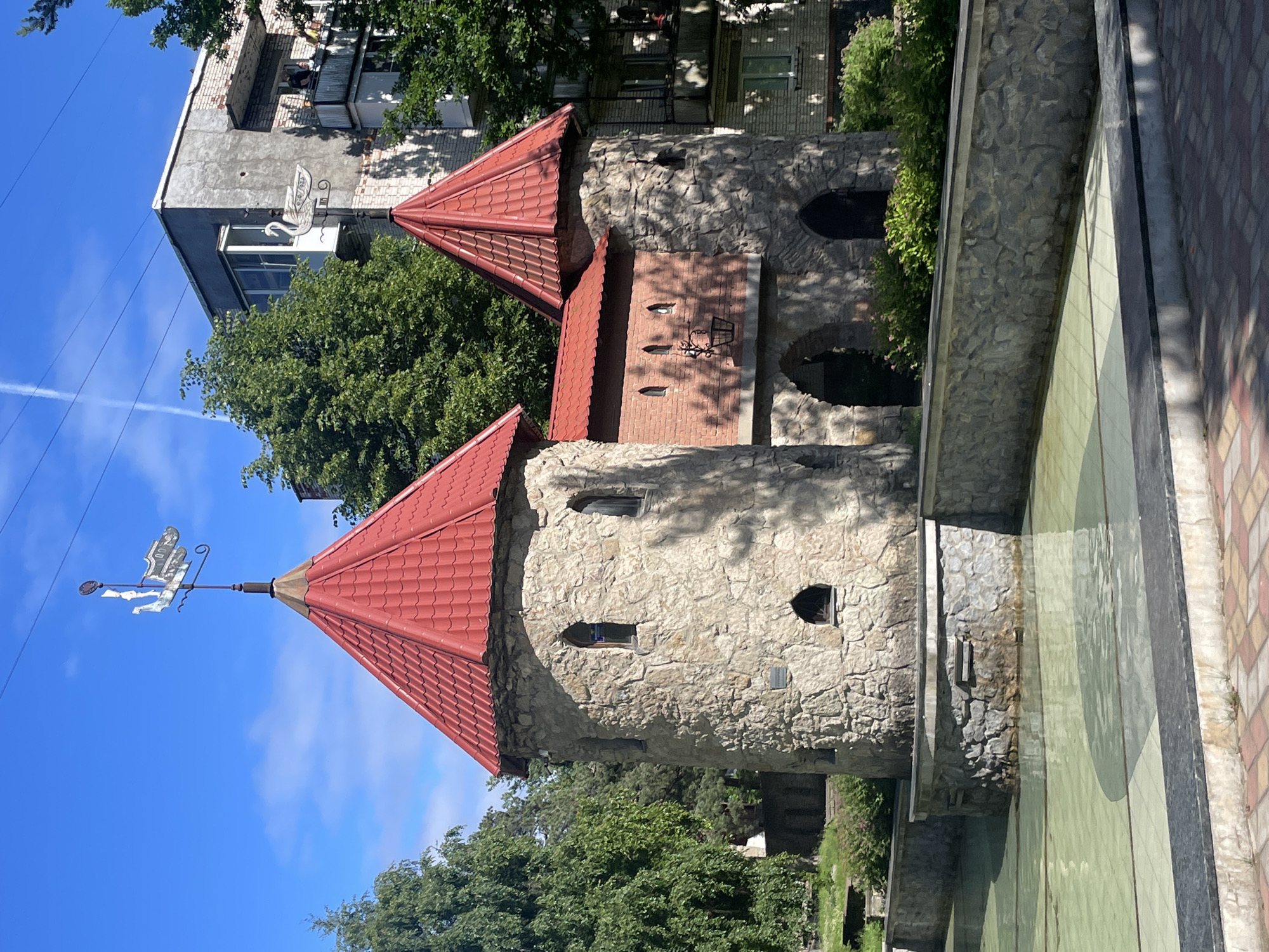 This is a vertical, eye-level, medium shot of a whimsical castle-like structure in Sosnoviy Bor, Russia. The structure features two conical red-tiled roofs and walls made of rough-hewn stone, with several small, dark arched windows. A weather vane shaped like a figure on a horse is mounted on the tip of the left turret, facing left. The castle appears to be part of a park or public space, as indicated by the adjacent paved walkway with a checkerboard pattern of pink and beige tiles, and a shallow, green-tiled pool or fountain in the foreground.
Behind the castle, an older, utilitarian apartment building with a gray facade and balconies is visible, partially obscured by lush green trees. The sky is a clear, bright blue with a few wispy white clouds and a faint contrail from an airplane. The lighting suggests it is daytime, likely midday or early afternoon, given the bright sunlight and distinct shadows.
There is no visible text in the image. The scene evokes a sense of playfulness and perhaps a touch of fairy tale fantasy, contrasting with the more mundane backdrop of the apartment building. The overall impression is a charming and unusual architectural feature set within an urban environment.