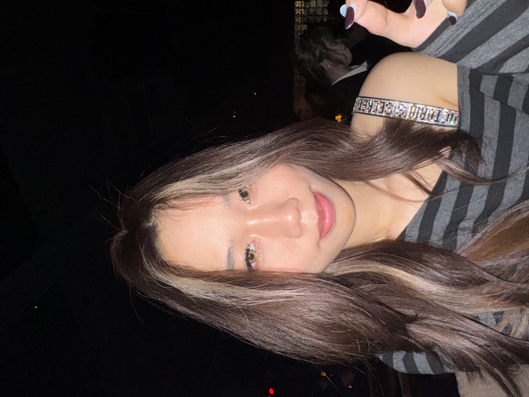 The image displays a young East Asian female in a dimly lit, nocturnal environment, located in Liangjiang, China. She occupies the left half of the frame, facing the camera with a subtle smile. Her long, dark brown hair, accentuated with prominent blonde highlights, extends over her shoulders. She wears a grey and black horizontally striped, off-the-shoulder top. A sparkling, rhinestone-embellished strap on her visible right shoulder bears the text "VICTORIA'S SECRET". Her right hand is partially visible on the far right, with fingernails painted dark red with light blue tips. In the blurred, dark background, a male figure is discernible, wearing a dark jacket over a white collared shirt, with his head slightly bowed. Distant, indistinct orange and red urban lights are visible further in the background, suggesting a city setting at night.