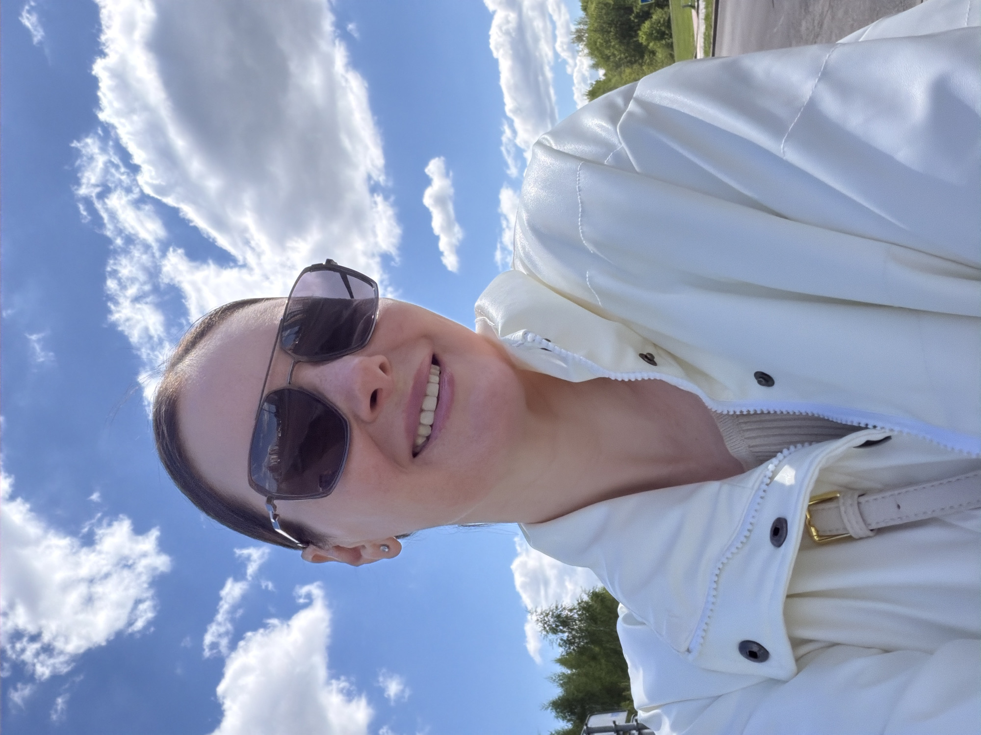 A woman is outdoors on a sunny day in an urban area. She is wearing sunglasses and a white jacket, with blue sky and scattered clouds visible in the background. Trees line a road in the distance.